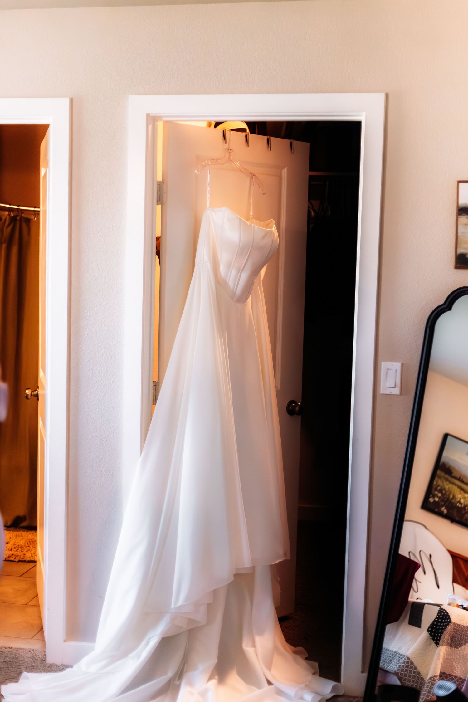 White wedding dress hanging in a doorway, with a flowing train pooling on the floor in a softly lit room.