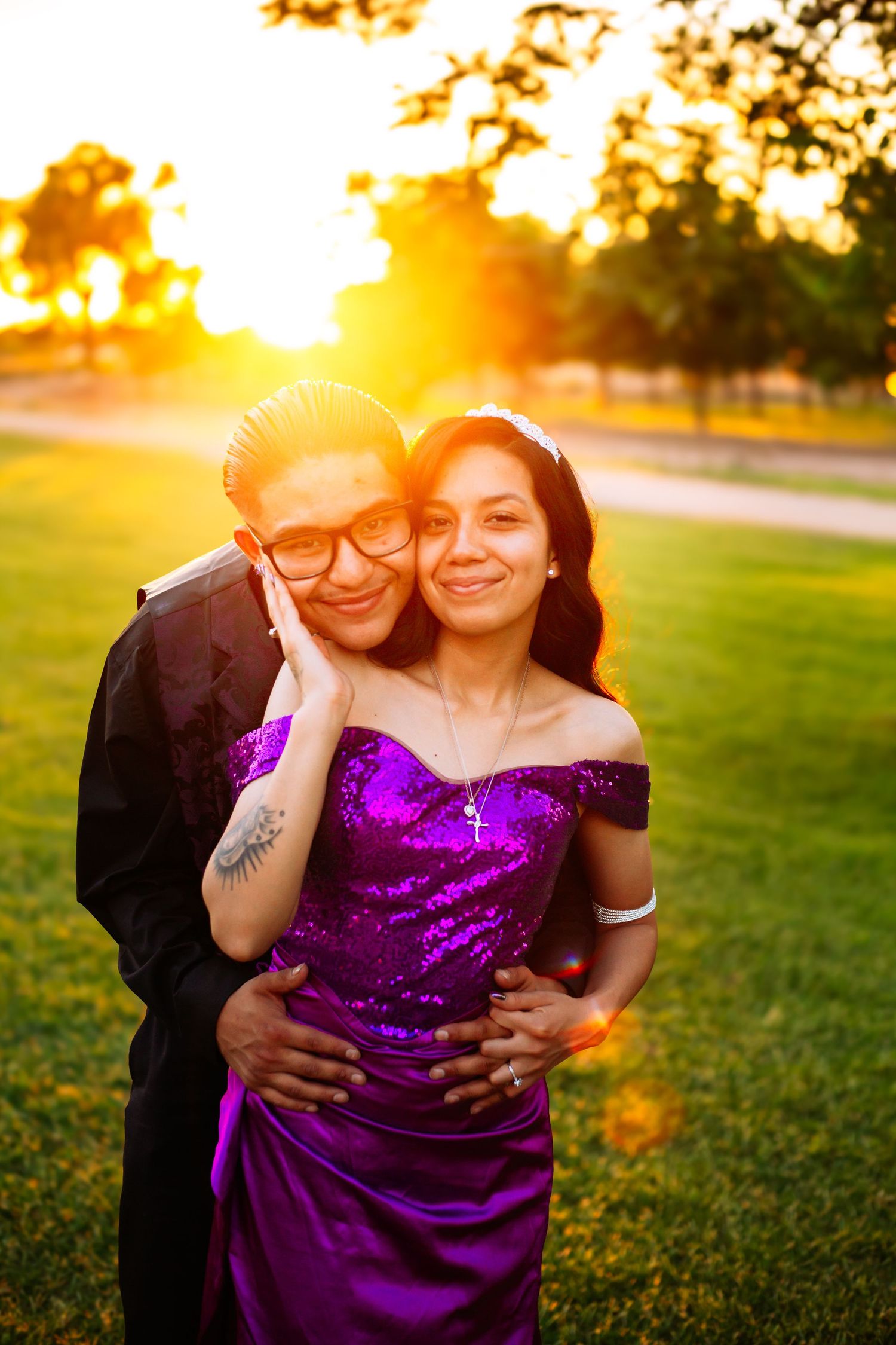 Couple posing at golden hour sunset in park, woman in purple sequin dress, man in black suit embracing her.