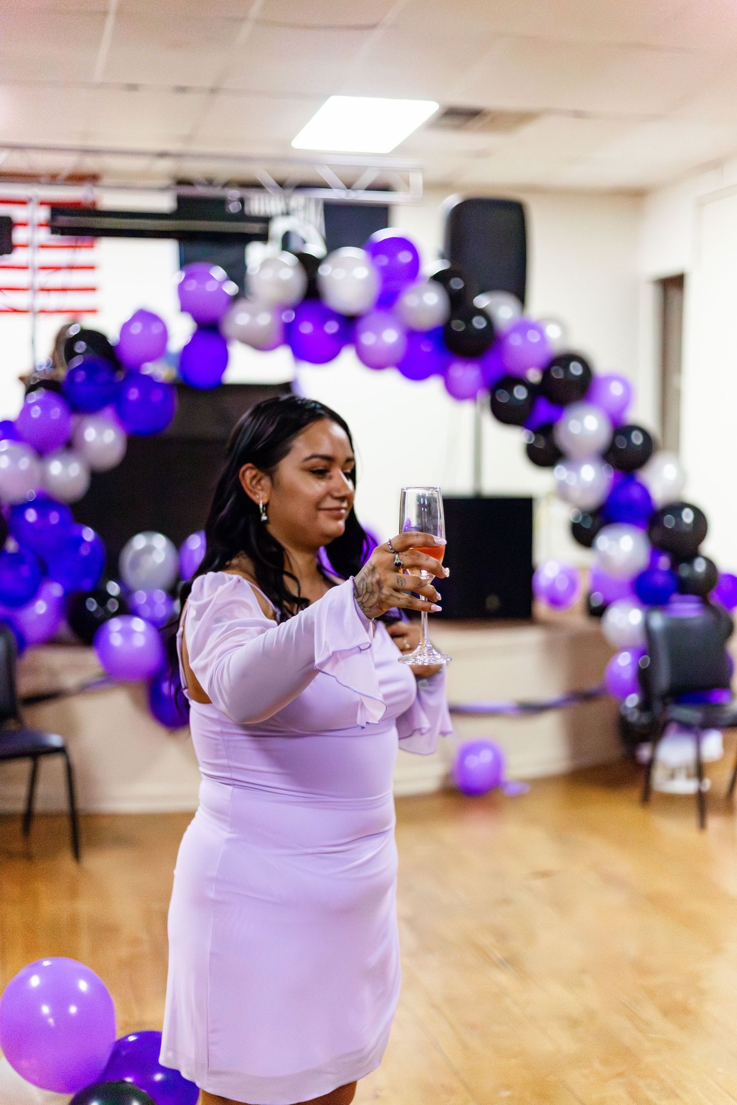 Woman in white dress toasting with champagne at a purple, black, and silver balloon arch party celebration.