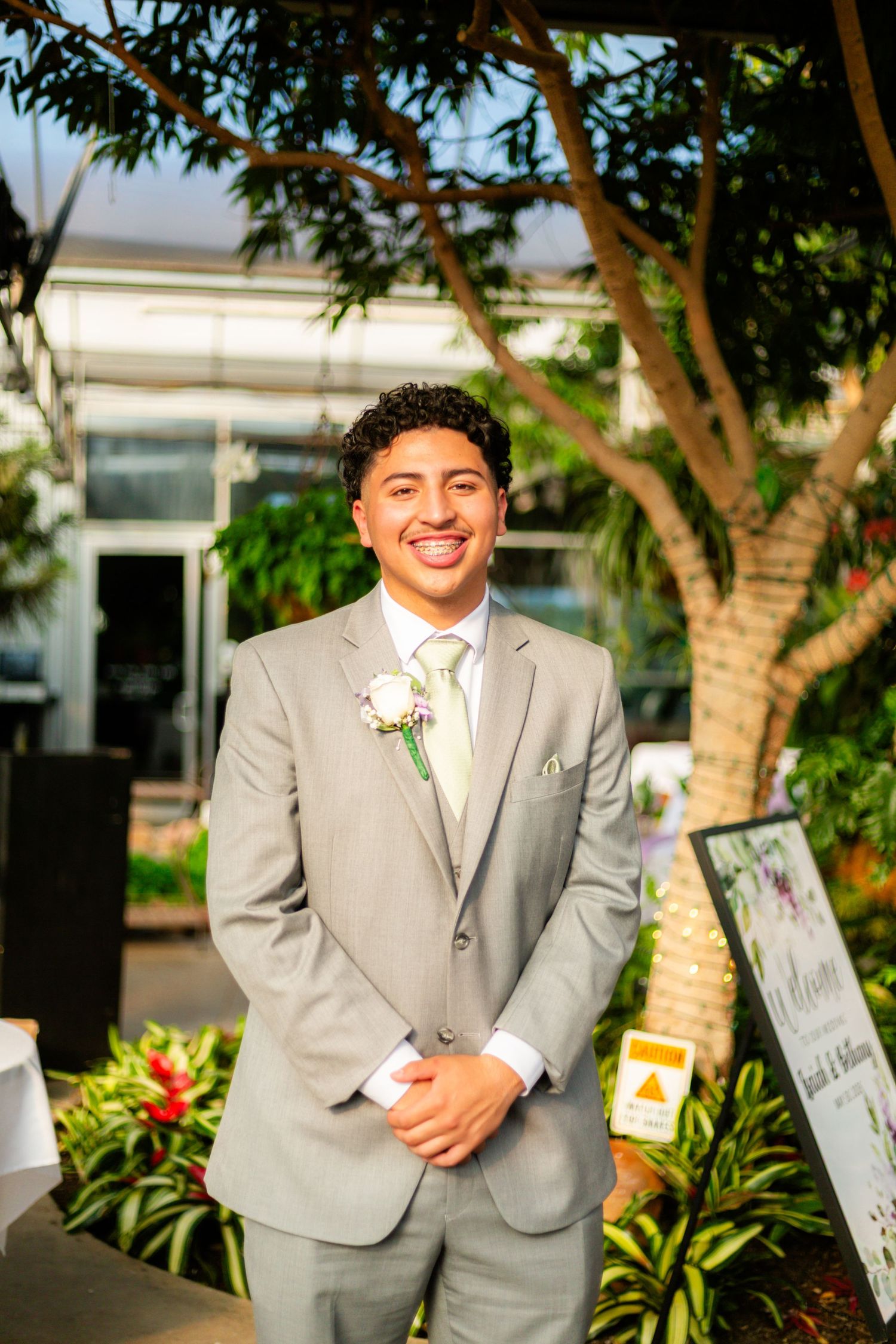 A smiling young man in a gray suit with white boutonniere stands outdoors at a wedding venue.