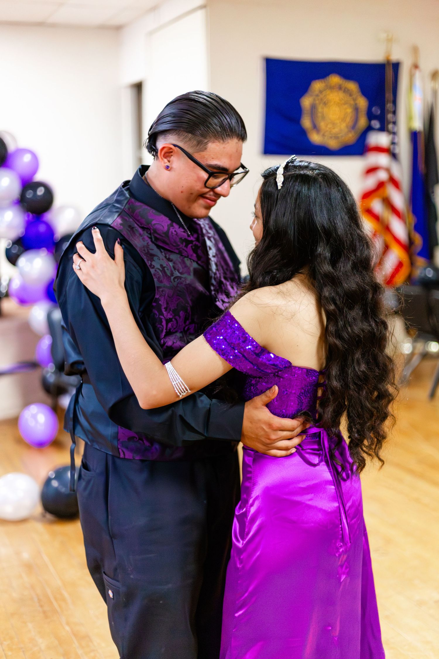 Couple sharing a dance at prom, wearing matching purple outfits, with balloons and flags in the background.
