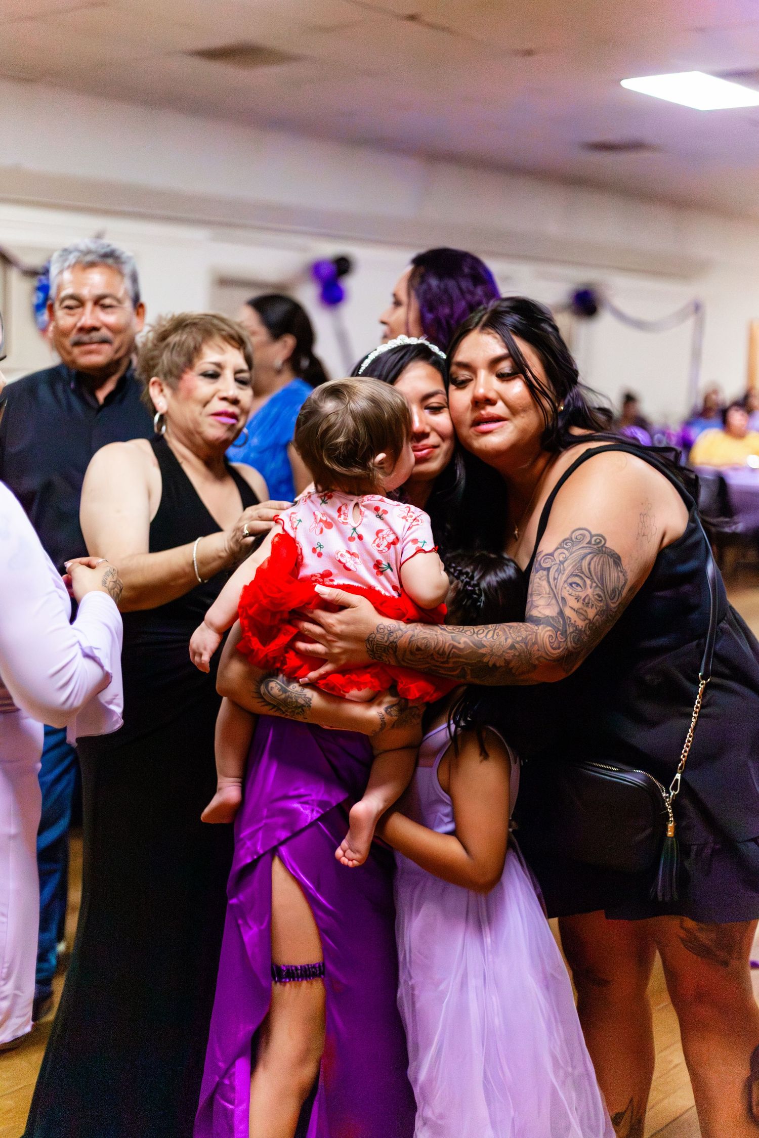Family and friends gather at a festive celebration, sharing a warm embrace with a baby in a red outfit.