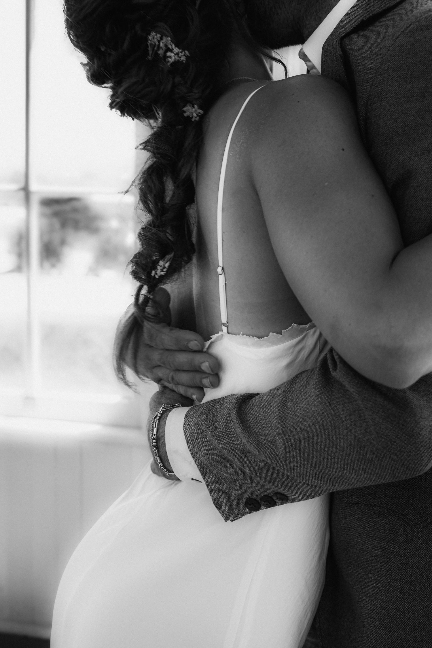 Black and white photo of a bride and groom embracing, showing her braided hair and wedding dress back detail.