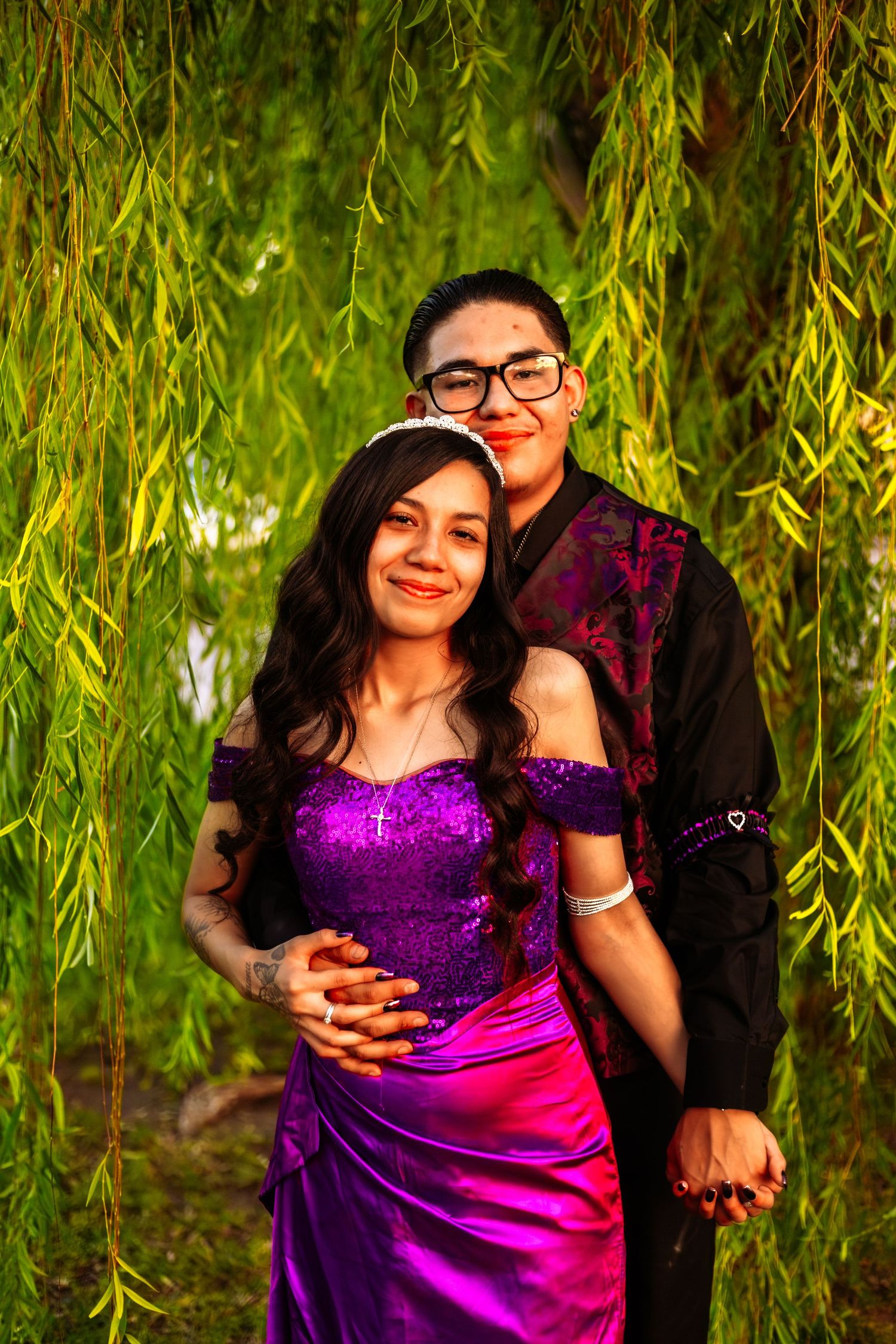 Couple posing under weeping willow, woman in purple sequin gown with tiara, man in black suit embracing her.