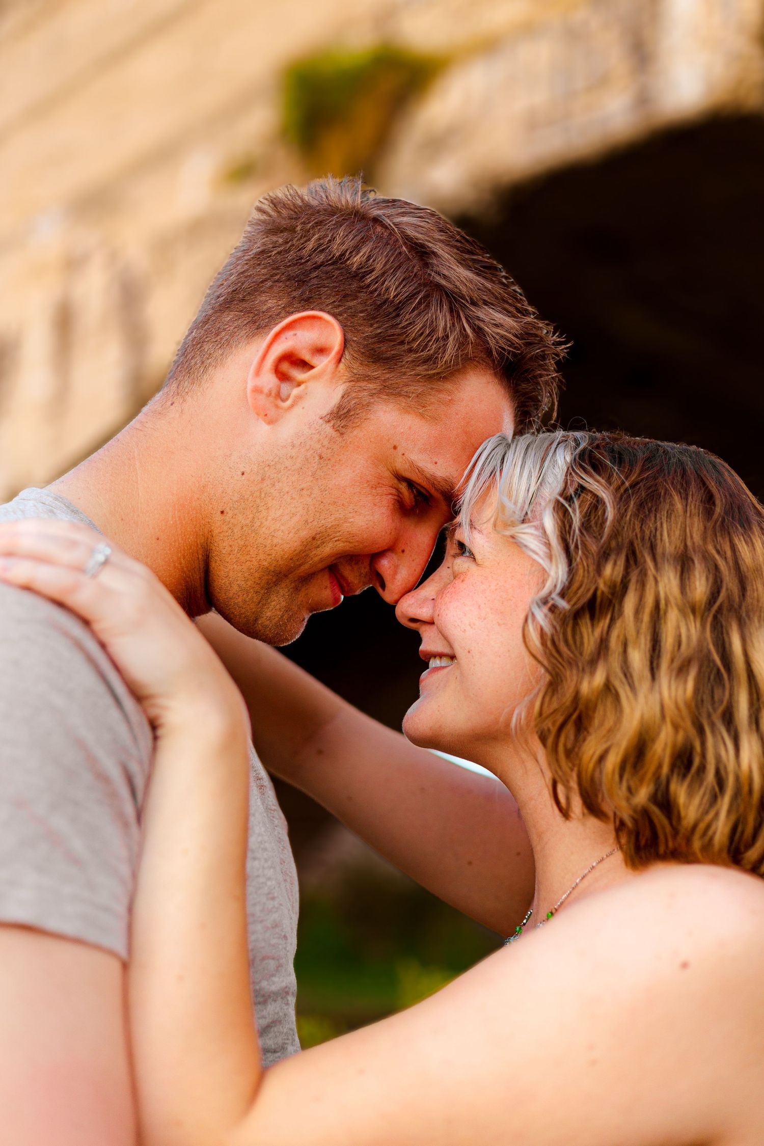 Romantic couple touching foreheads and smiling outdoors near a stone arch in warm golden light.