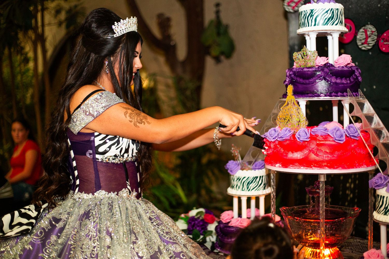 A young woman in a purple quinceañera gown and tiara cuts a colorful multi-tiered celebration cake at her party.