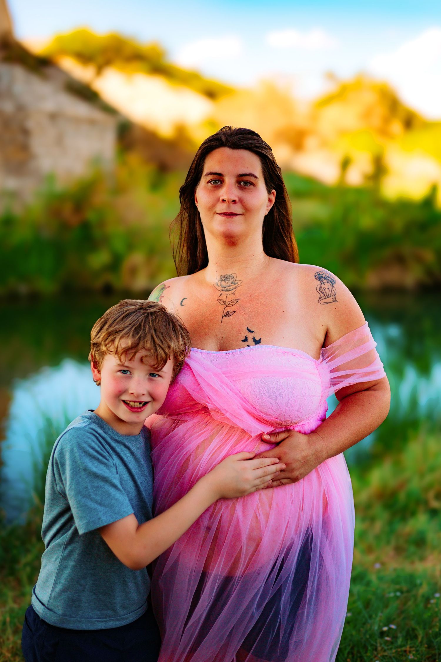 Pregnant woman in pink tulle dress with tattoos hugs smiling young boy outdoors near a pond at golden hour.
