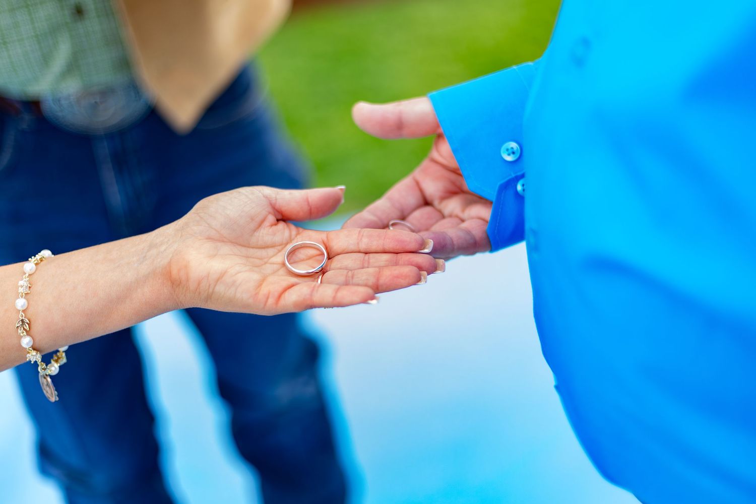 A close-up of two hands exchanging a gold wedding ring outdoors, symbolizing love and commitment.