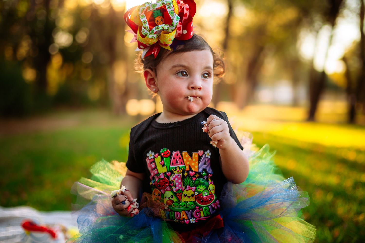 Toddler girl in colorful tutu skirt and birthday shirt with festive crown headband outdoors.