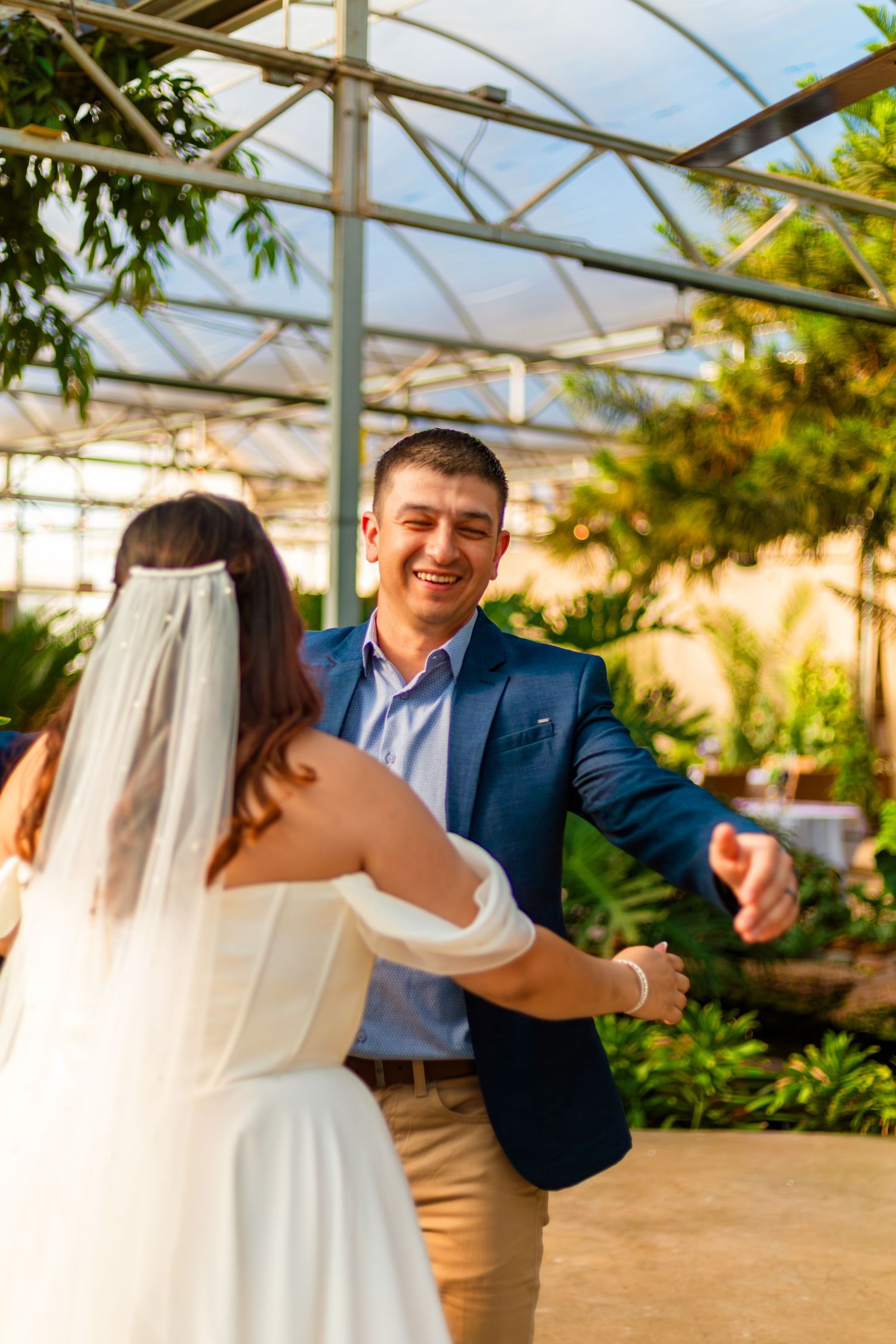 Smiling groom in blue suit dancing with bride in white dress and veil inside a lush greenhouse venue.