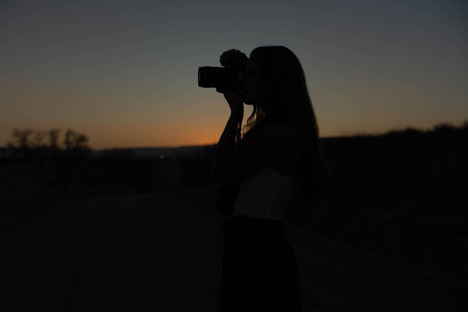 Silhouette of a woman photographer shooting with a DSLR camera against a colorful sunset sky.