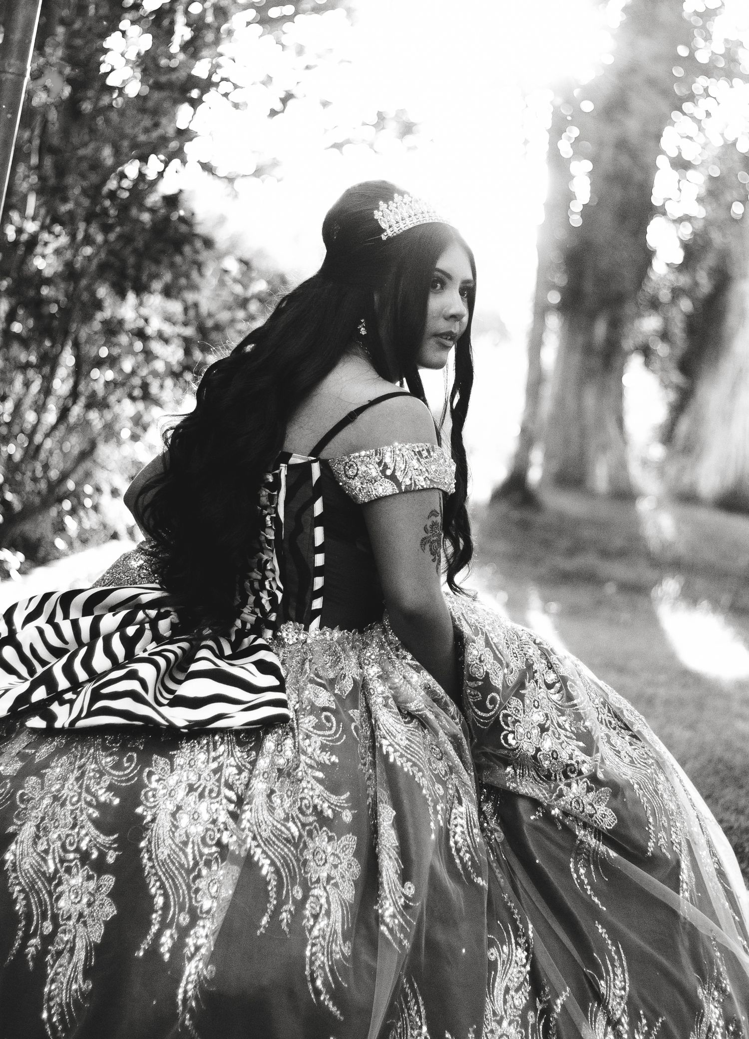 Black and white photo of a young woman in an ornate off-shoulder ballgown with a tiara, posed outdoors.