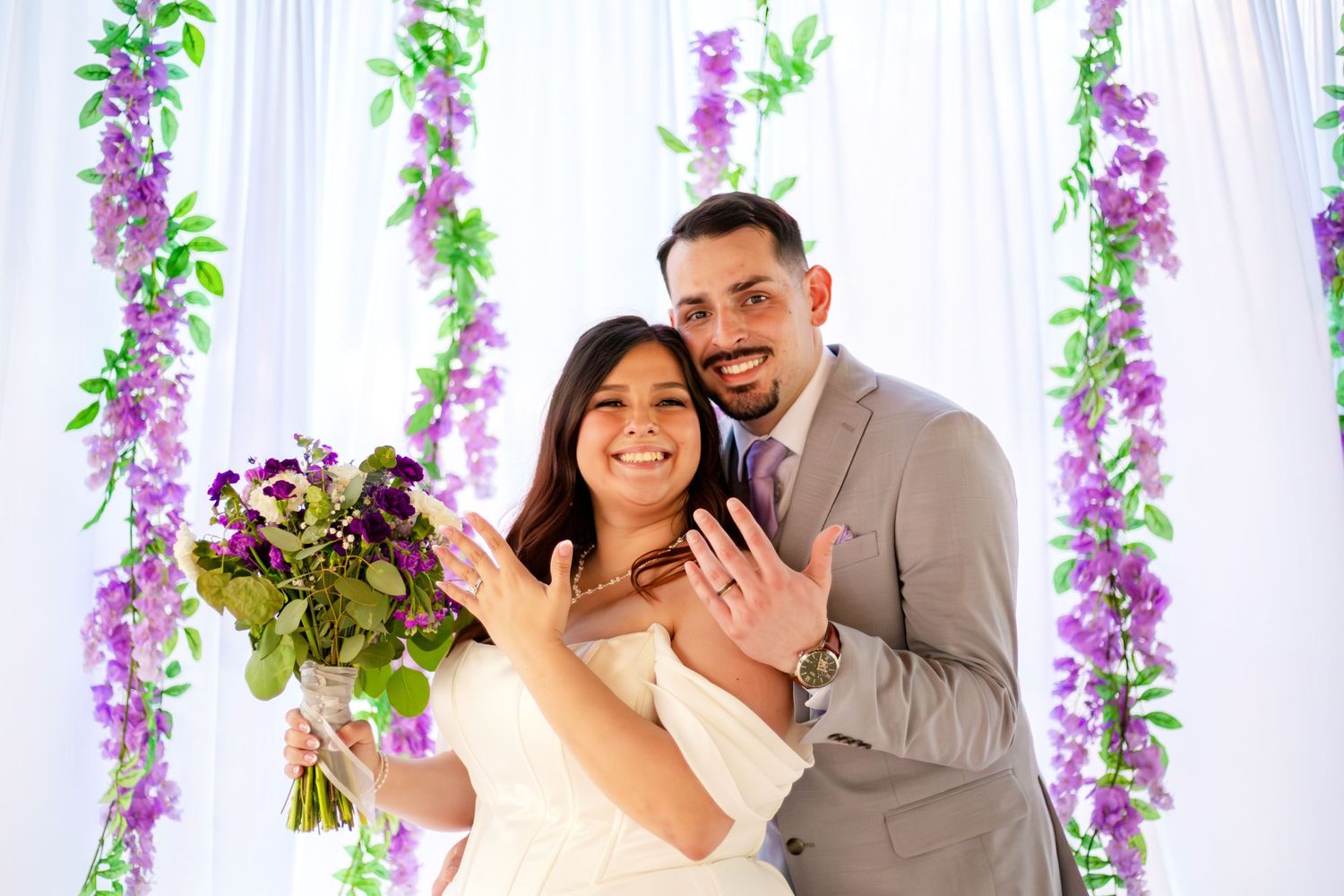 Smiling newlyweds pose before purple floral arch, bride holding bouquet, groom in gray suit, both showing wedding rings.