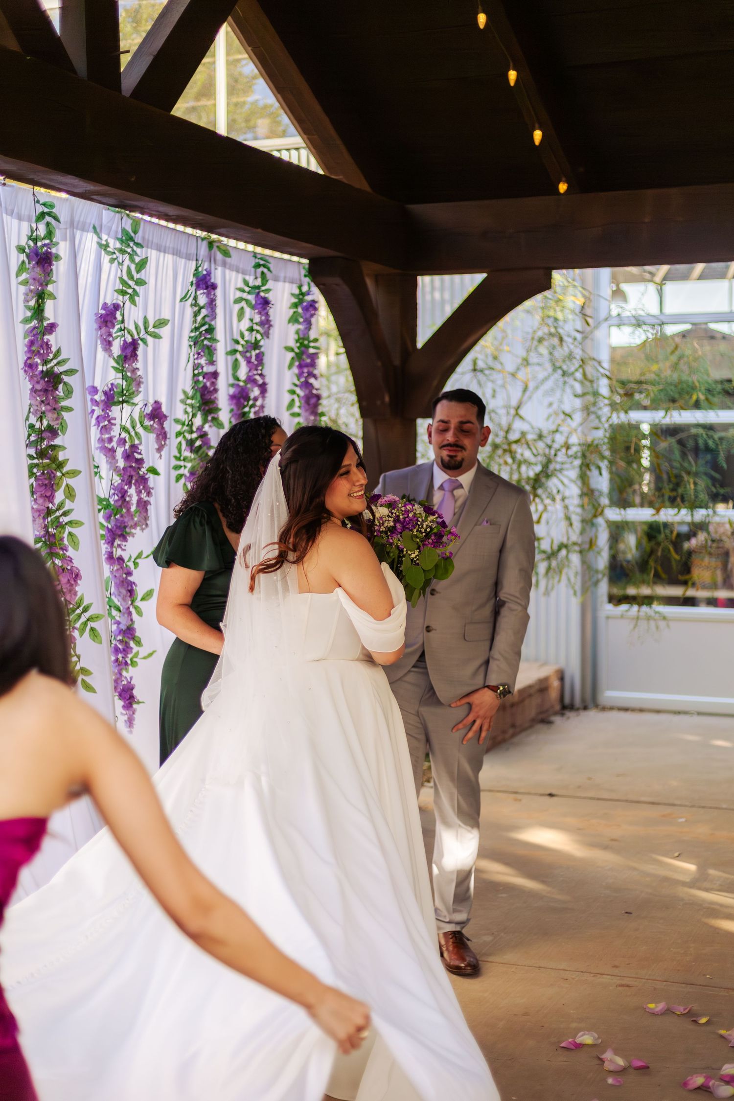 Bride in white gown walks toward groom at outdoor wedding ceremony with purple floral backdrop.
