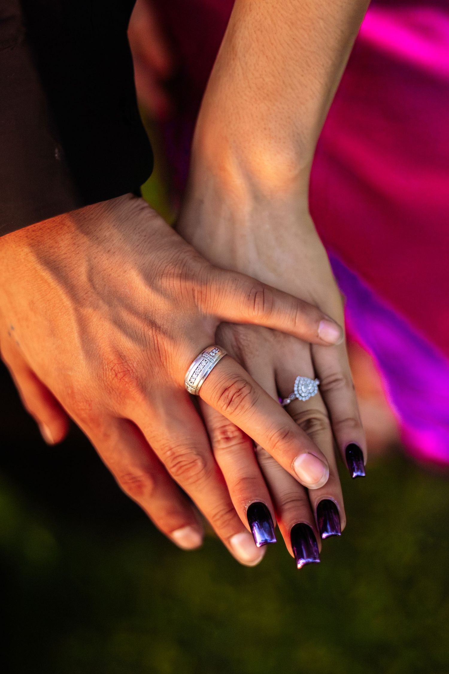 Close-up of couple's hands showing wedding rings, woman with dark purple nails and diamond engagement ring.