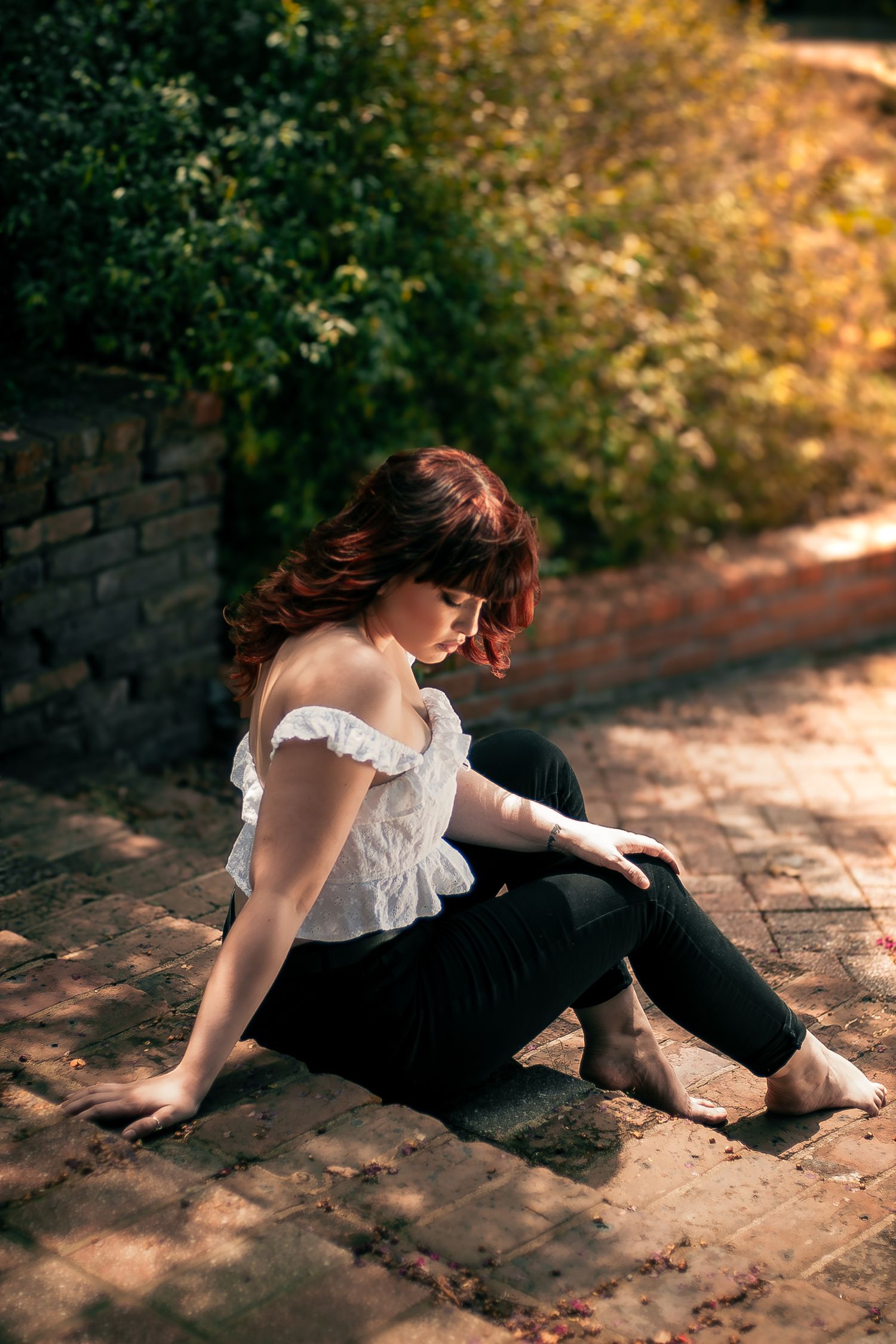 Redhead woman in white off-shoulder top and black pants sitting on brick path.