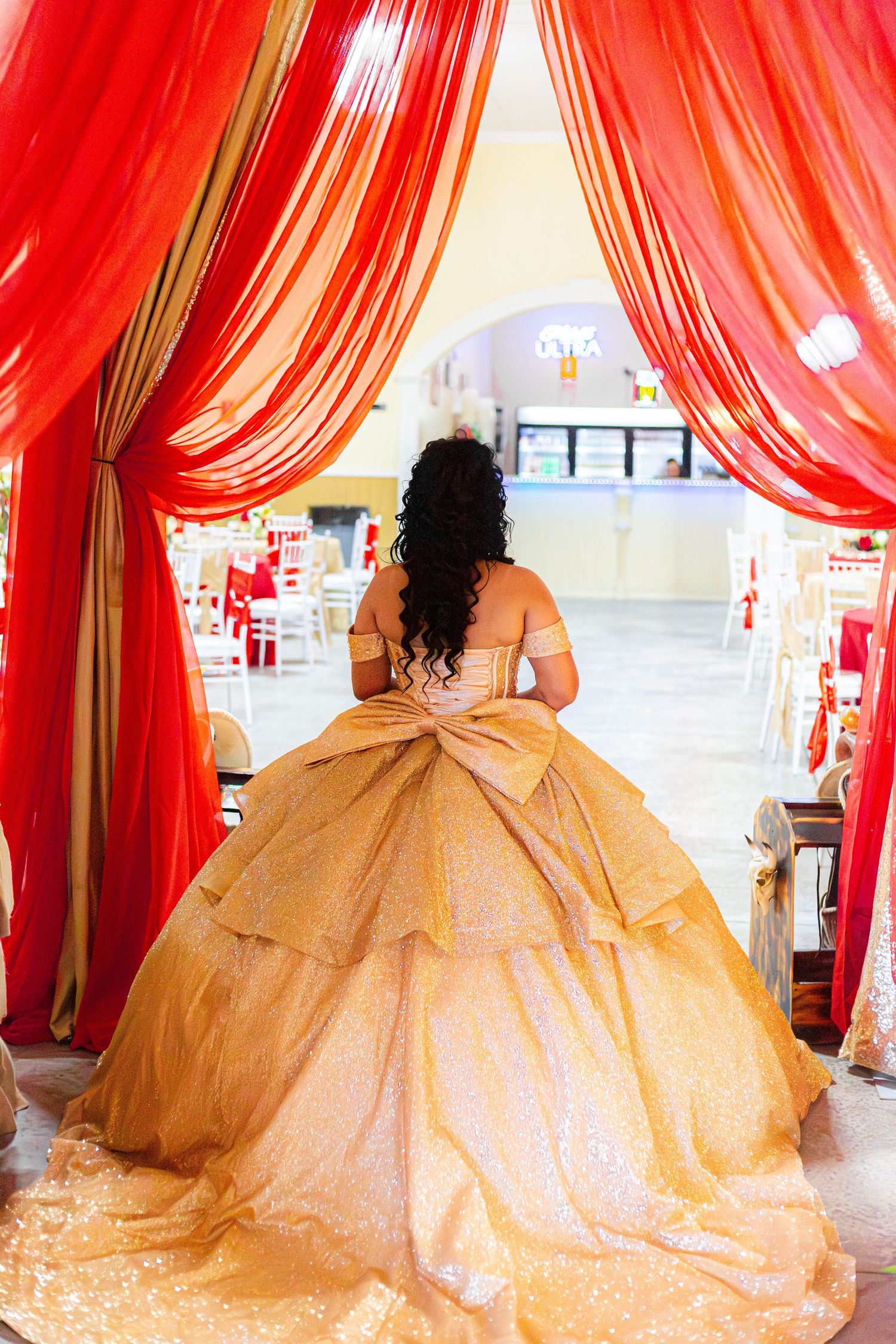 Young woman in gold quinceañera ball gown stands at red draped entrance of decorated celebration venue.