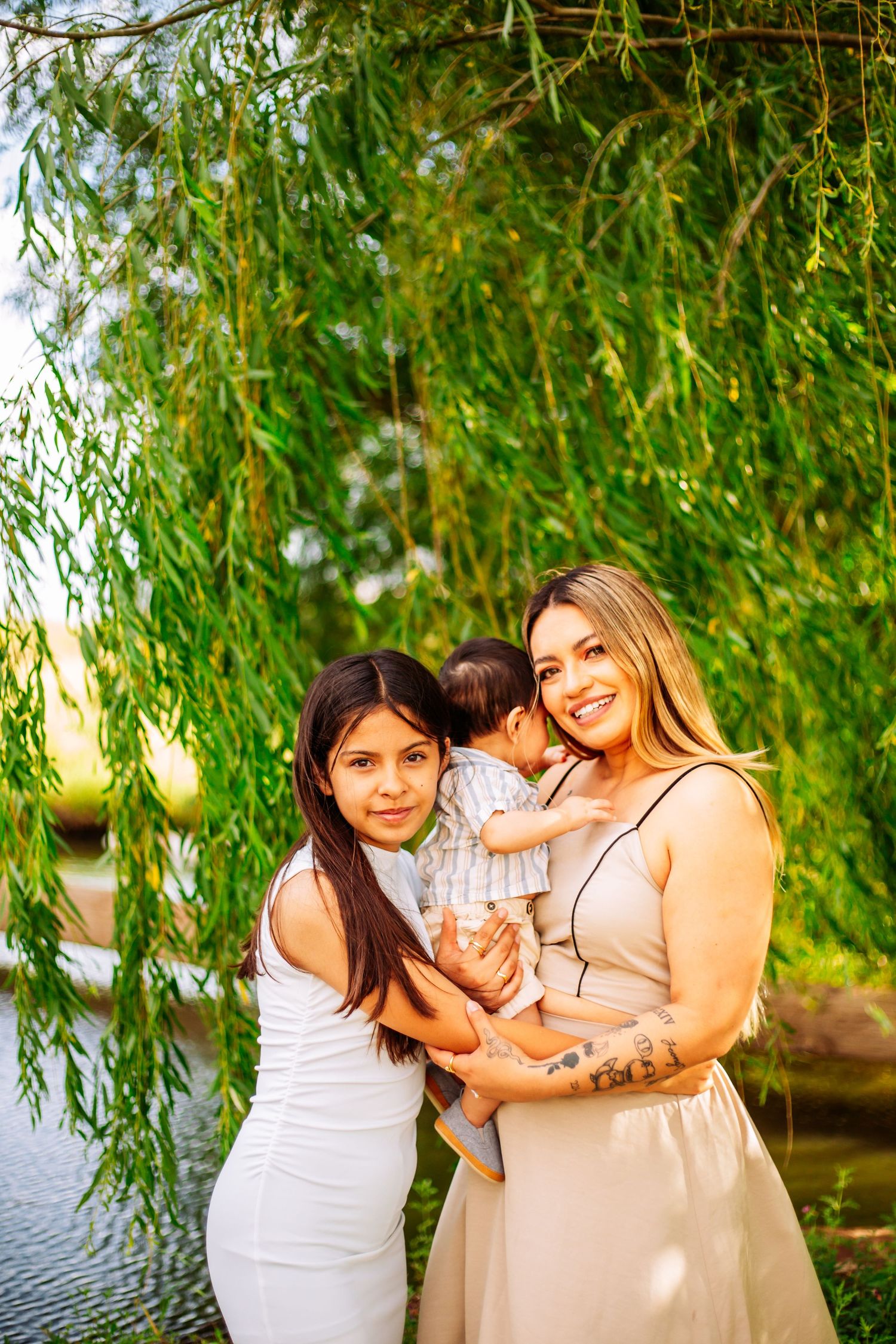 A mother and two children pose outdoors under a weeping willow tree near a pond on a sunny day.