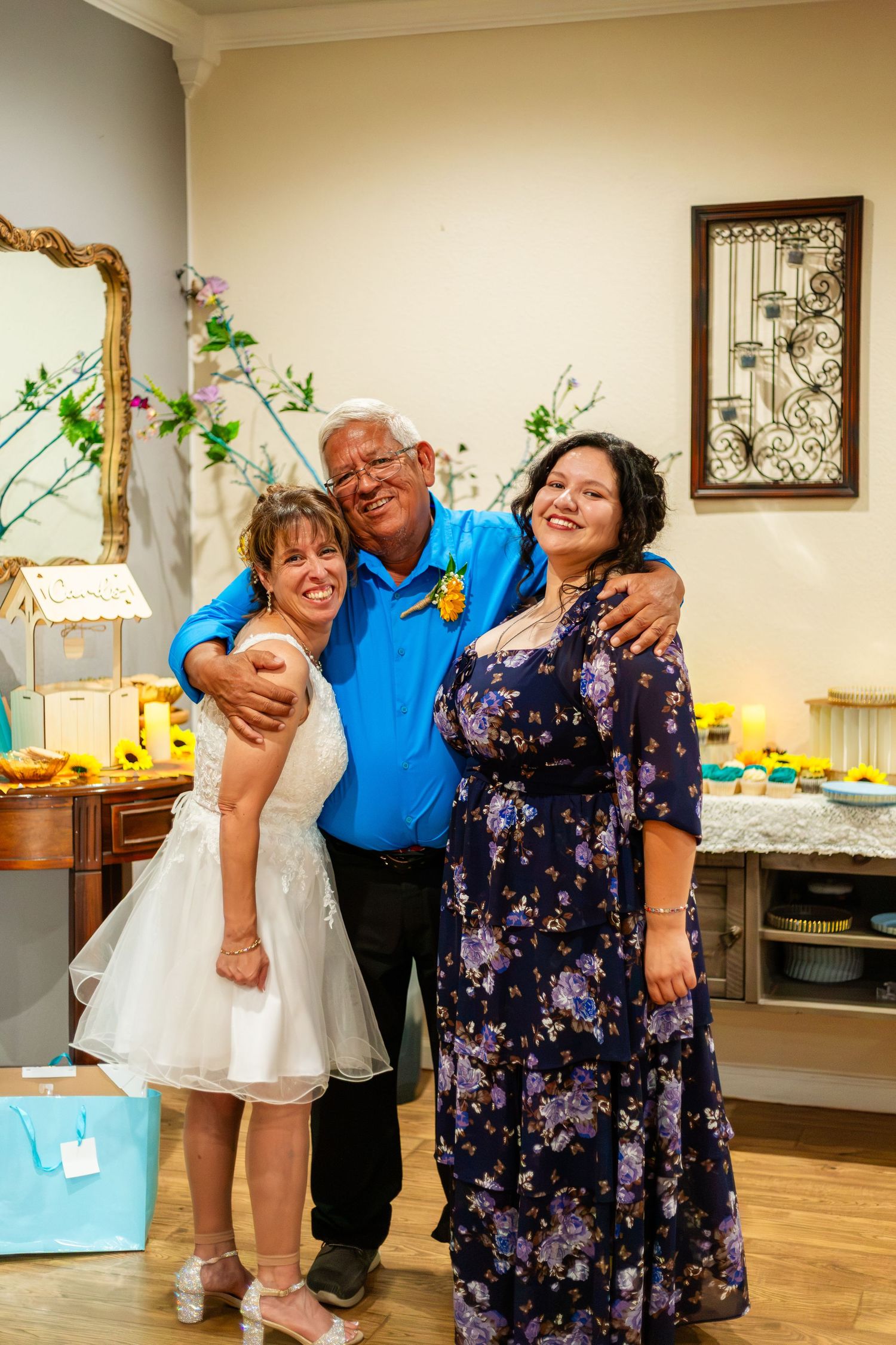 Three people smiling and posing together at a wedding celebration, with a decorated dessert table in the background.