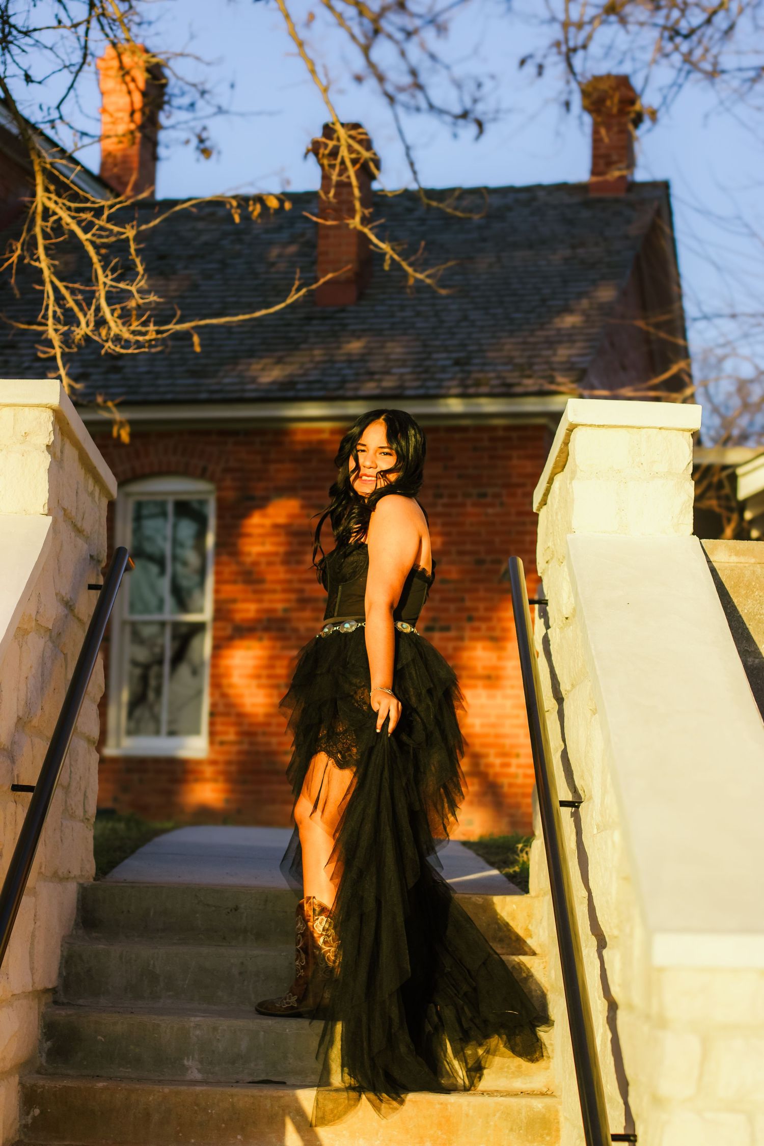 Woman in black tulle gown posing on stone steps outside brick building.