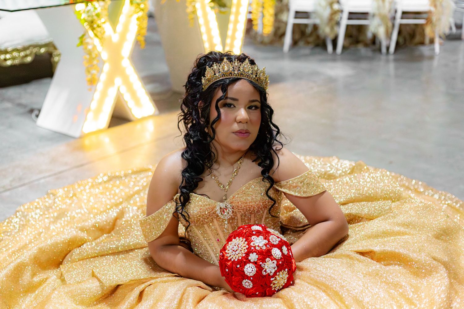 Young woman in gold quinceañera gown with crown holds red bouquet, posing elegantly at celebration venue.
