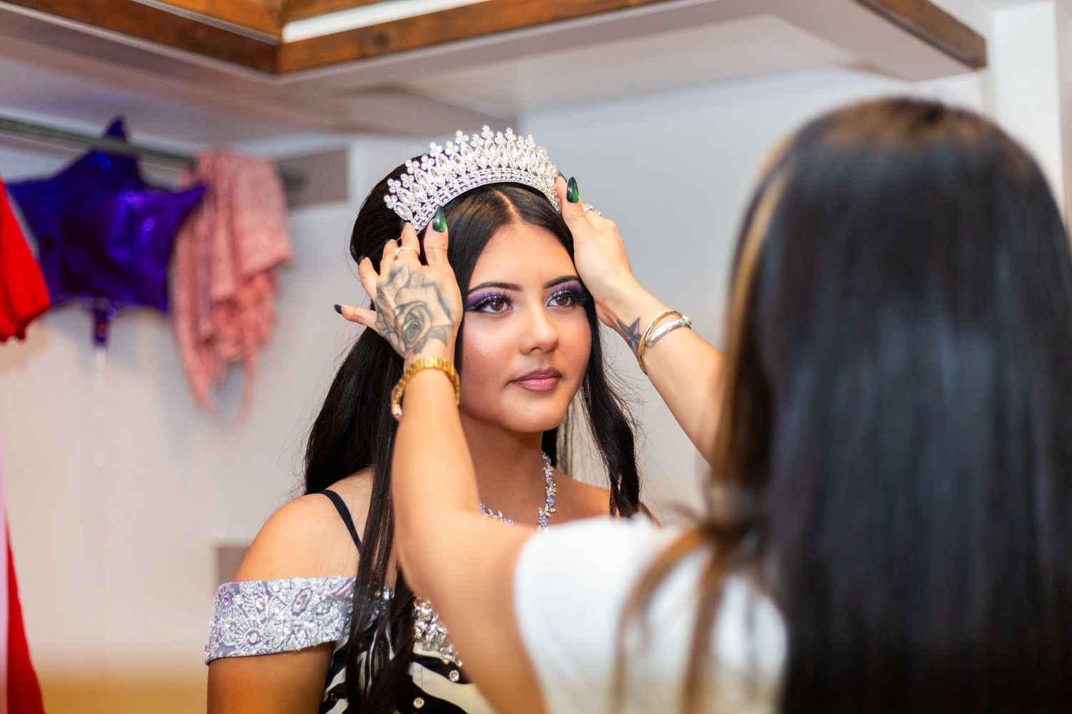 A woman places a sparkling tiara crown on a young woman in a silver dress, reflecting in a mirror.