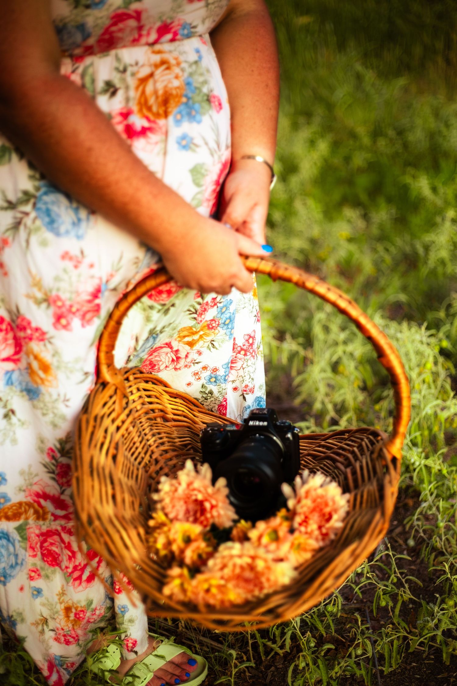Woman in floral dress holding wicker basket with Nikon camera and yellow flowers outdoors.