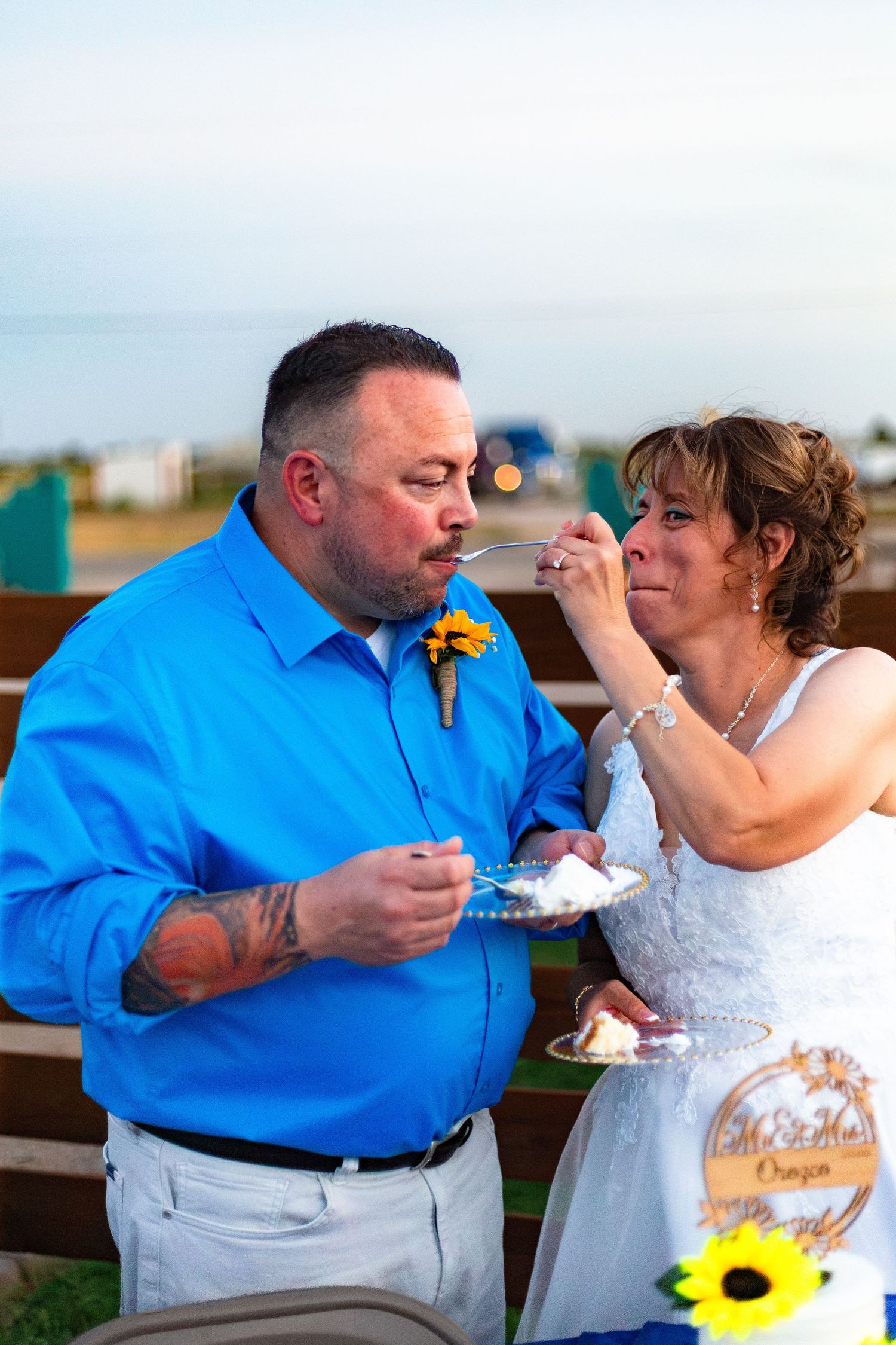 Bride feeds groom wedding cake at outdoor reception, both smiling and wearing sunflower accents.