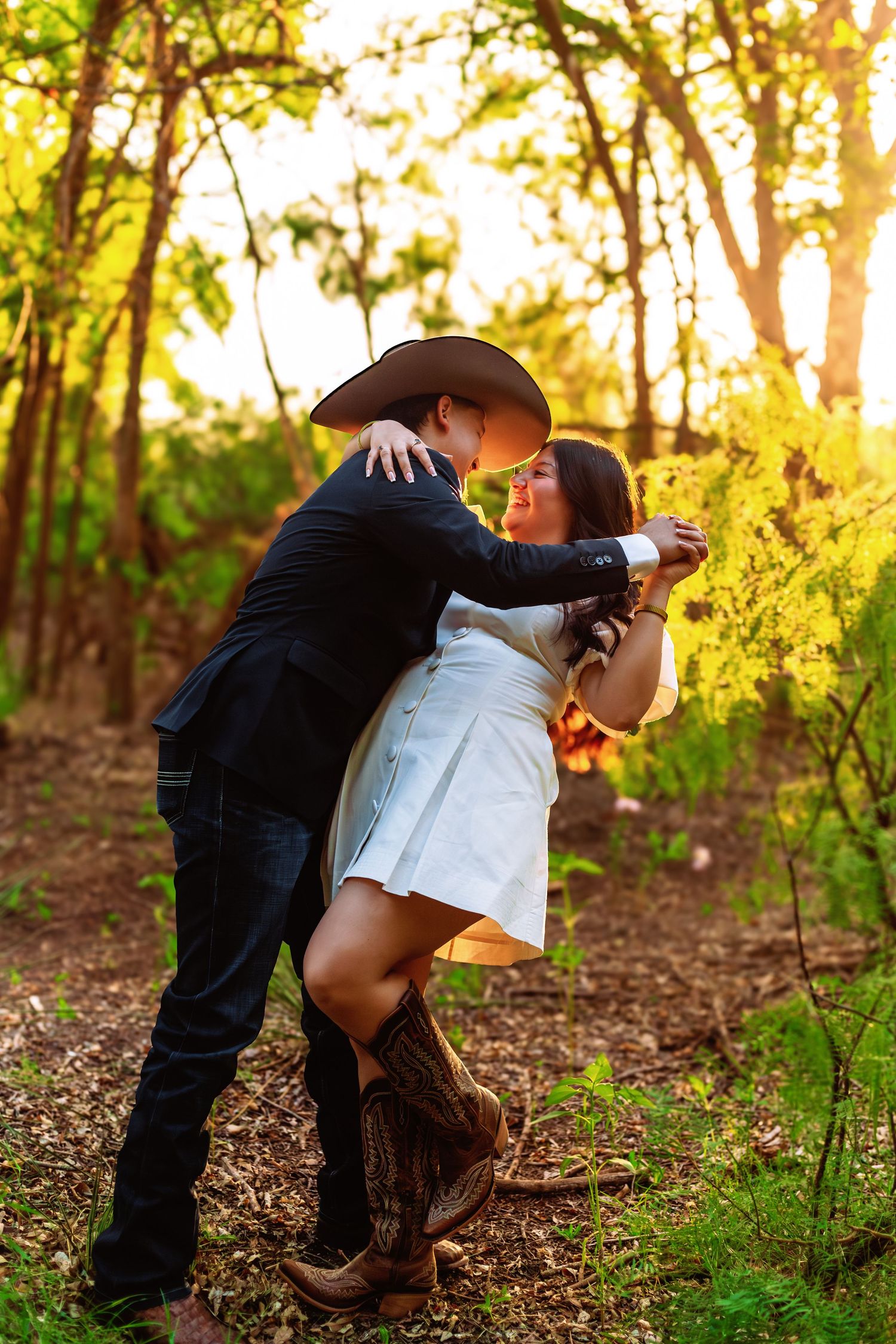 Couple dancing romantically in sunlit forest, man in cowboy hat and black jacket, woman in white dress and cowboy boots.