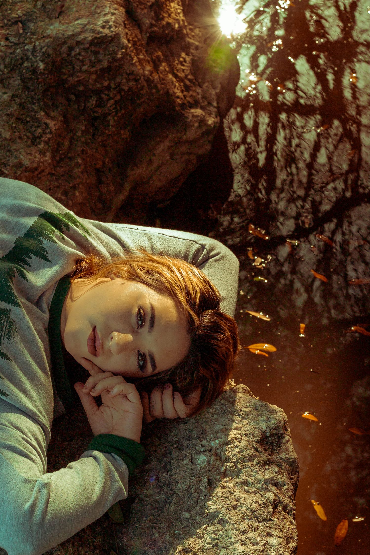 Young boy resting on a rock near water with autumn leaves in warm sunlight.