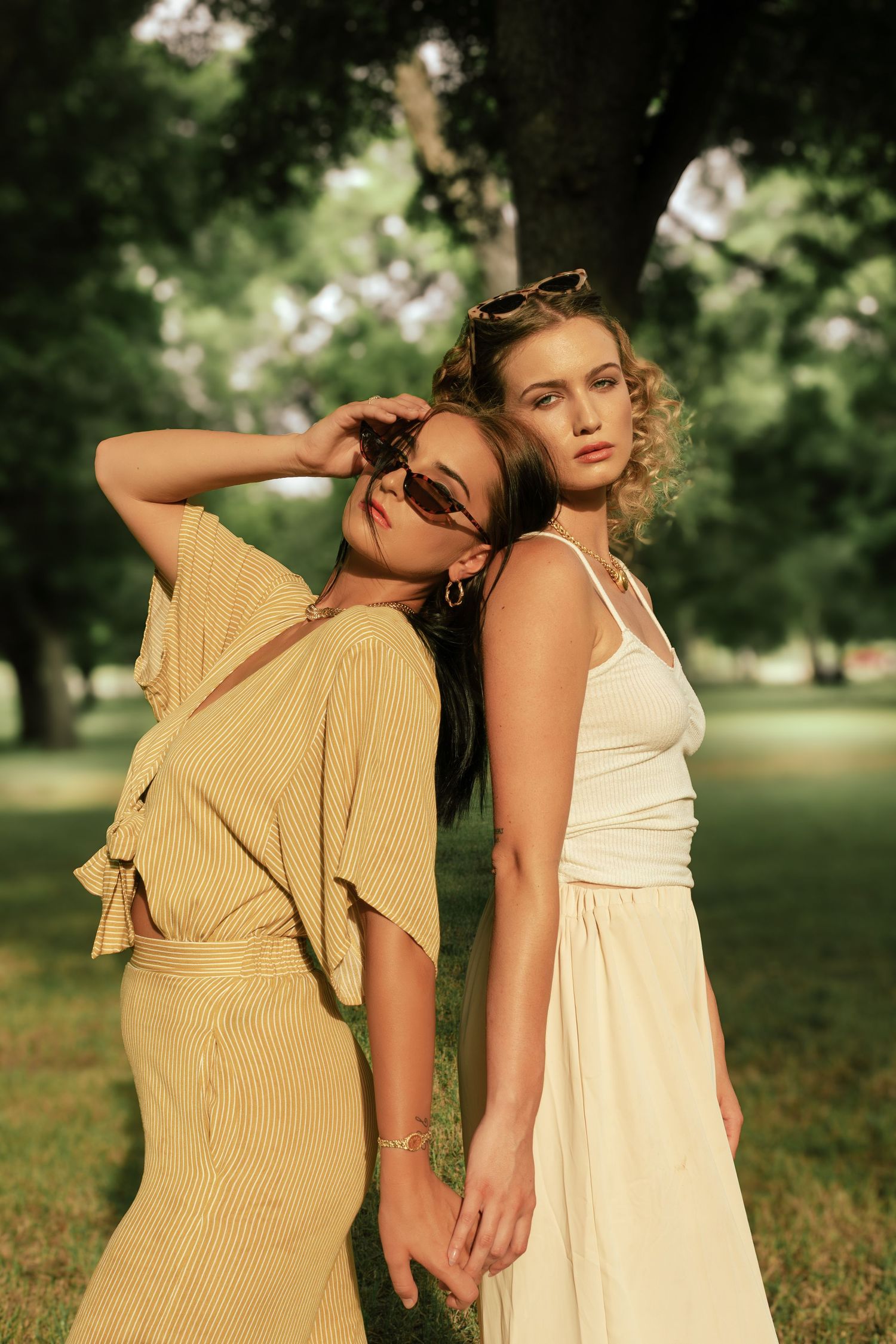 Two stylish women pose in a sunny park, one in yellow striped dress with sunglasses, other in cream sundress.