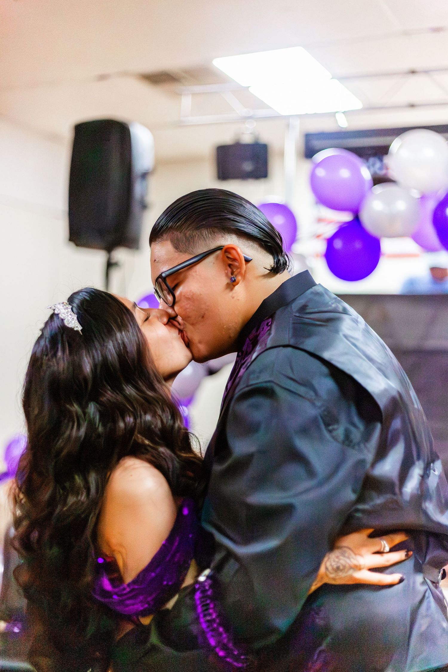 Couple sharing a romantic kiss at a purple-themed celebration with balloons in the background.
