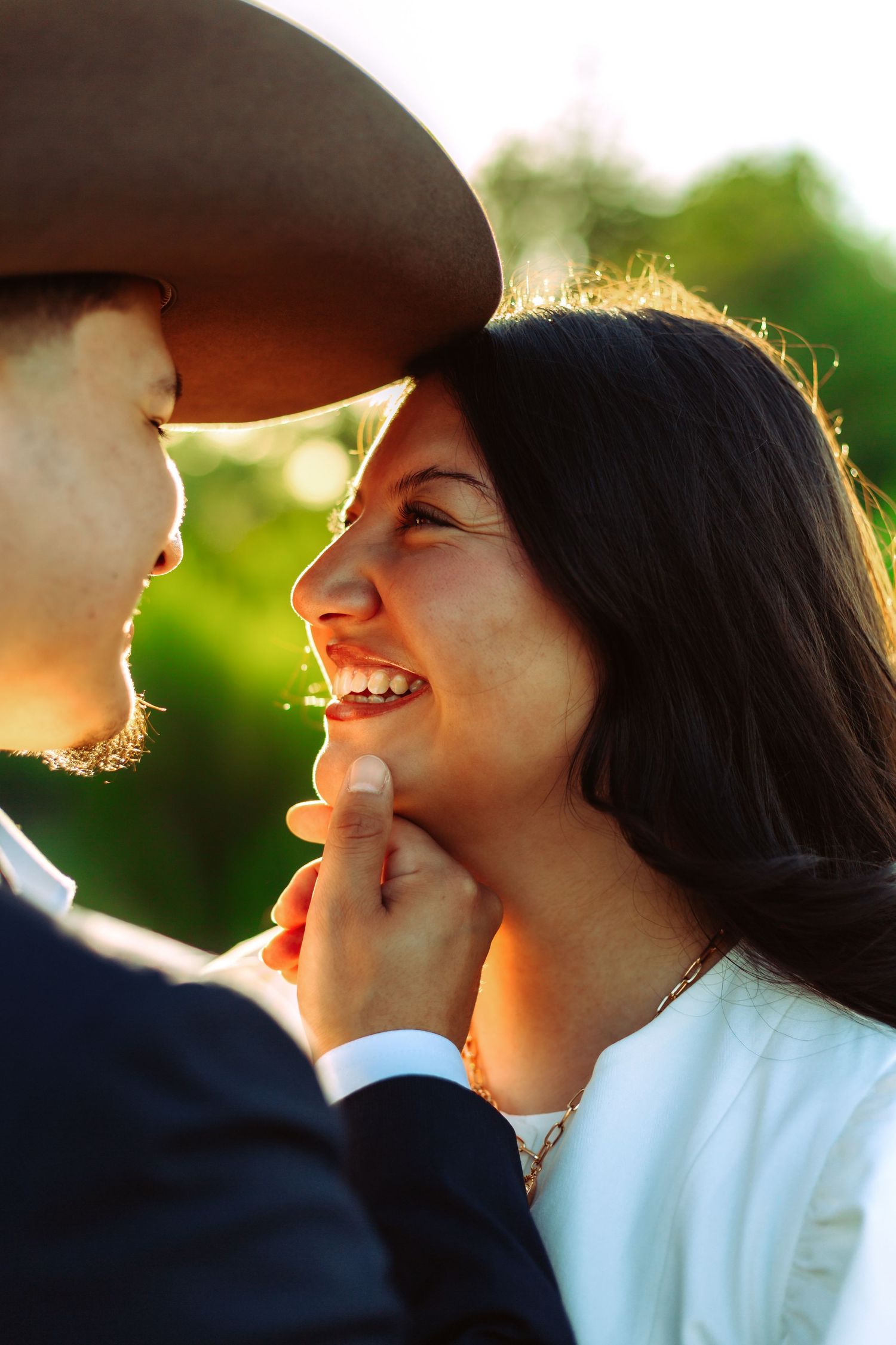 A man in a cowboy hat tenderly holds a smiling woman's chin in a romantic outdoor sunset portrait.