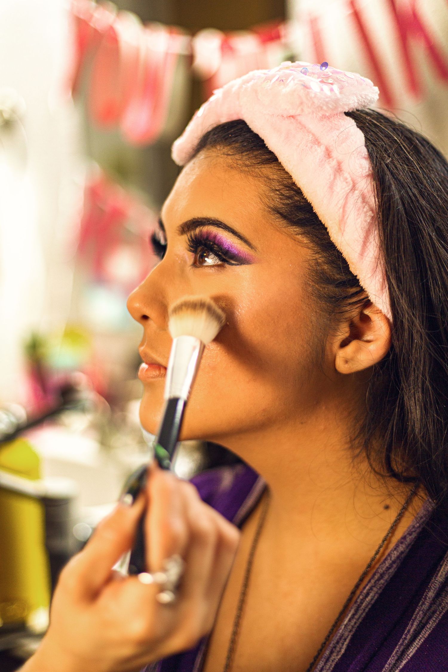 Young woman with pink headband getting makeup applied with brush, featuring purple eye shadow in vanity mirror setting.