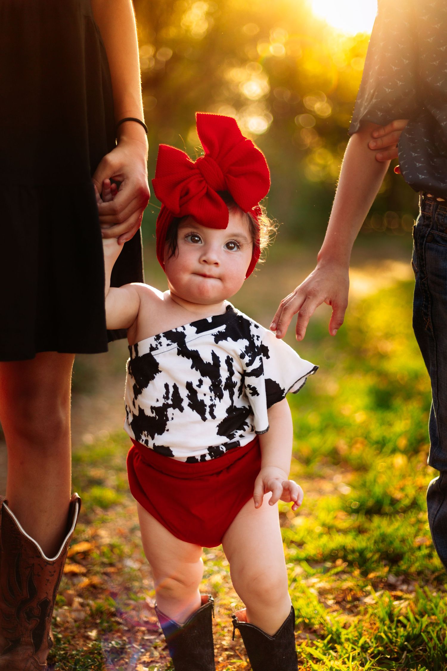 Adorable baby girl in cow print romper with red bow headband stands outdoors at golden hour held by parents.