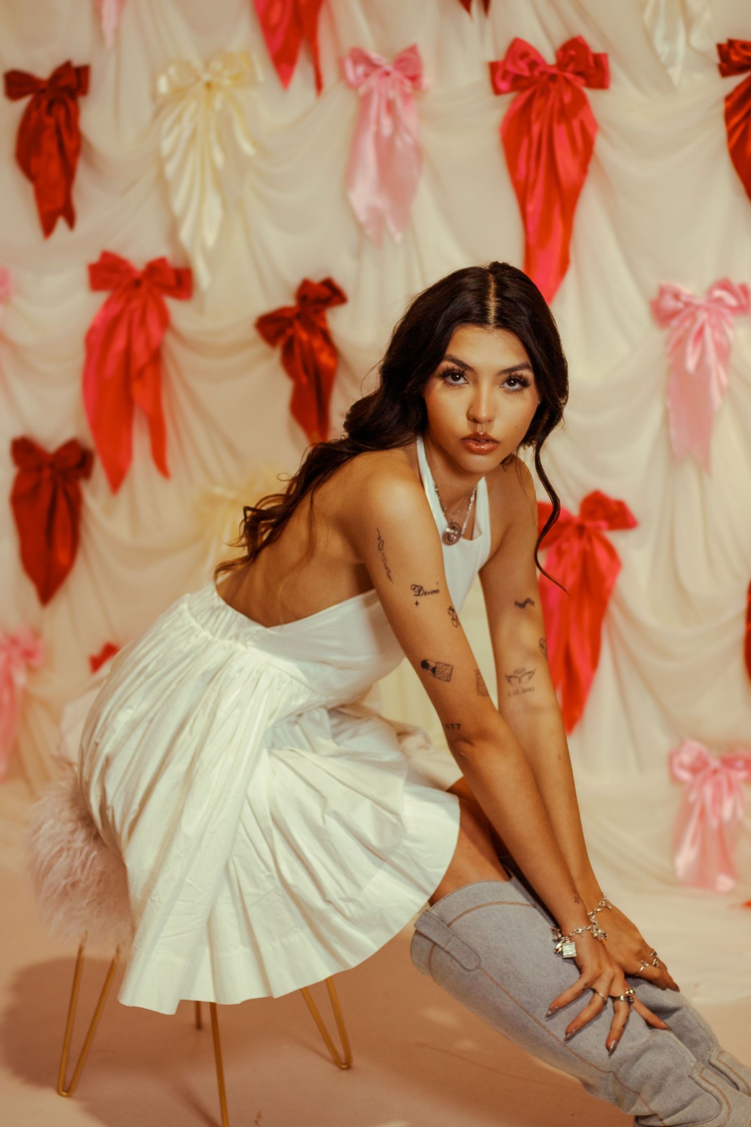 Tattooed woman in white halter dress posing before colorful bow-adorned backdrop.