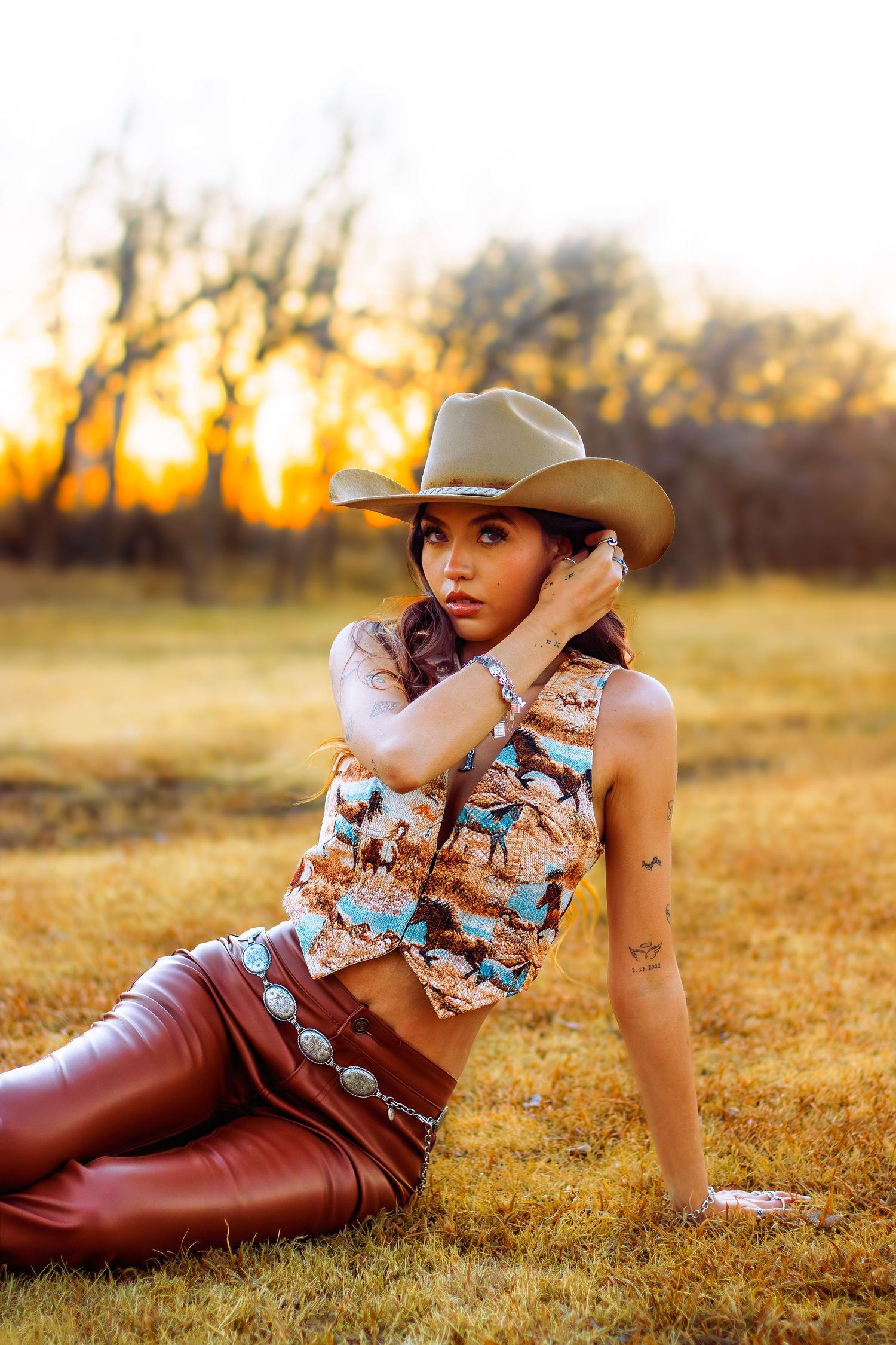 Woman in cowboy hat, patterned western top, leather pants poses in golden sunset field.