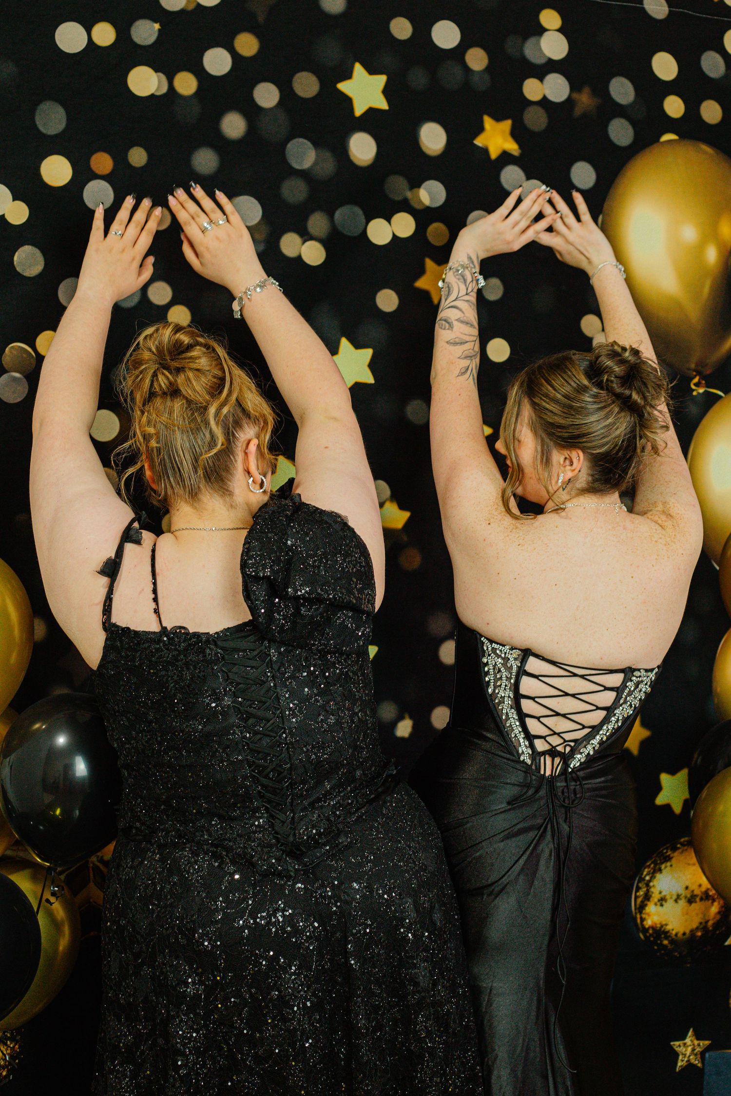 Two women in black formal gowns pose with arms raised at a glamorous party with gold balloons and star decorations.