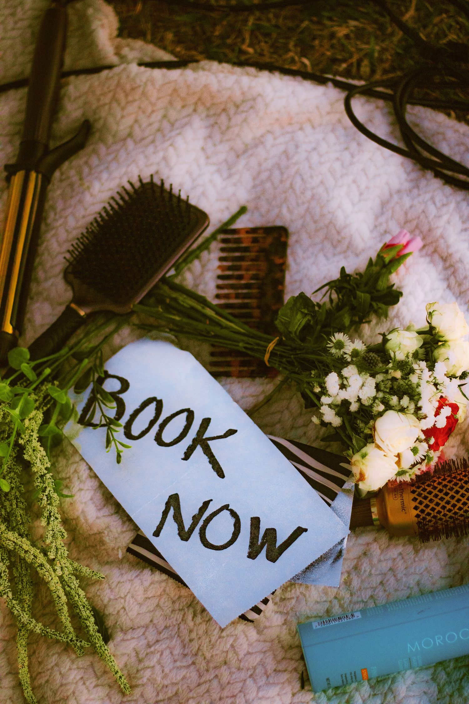 Flat lay of hair styling tools, flowers, combs, and a Book Now sign on white towel.