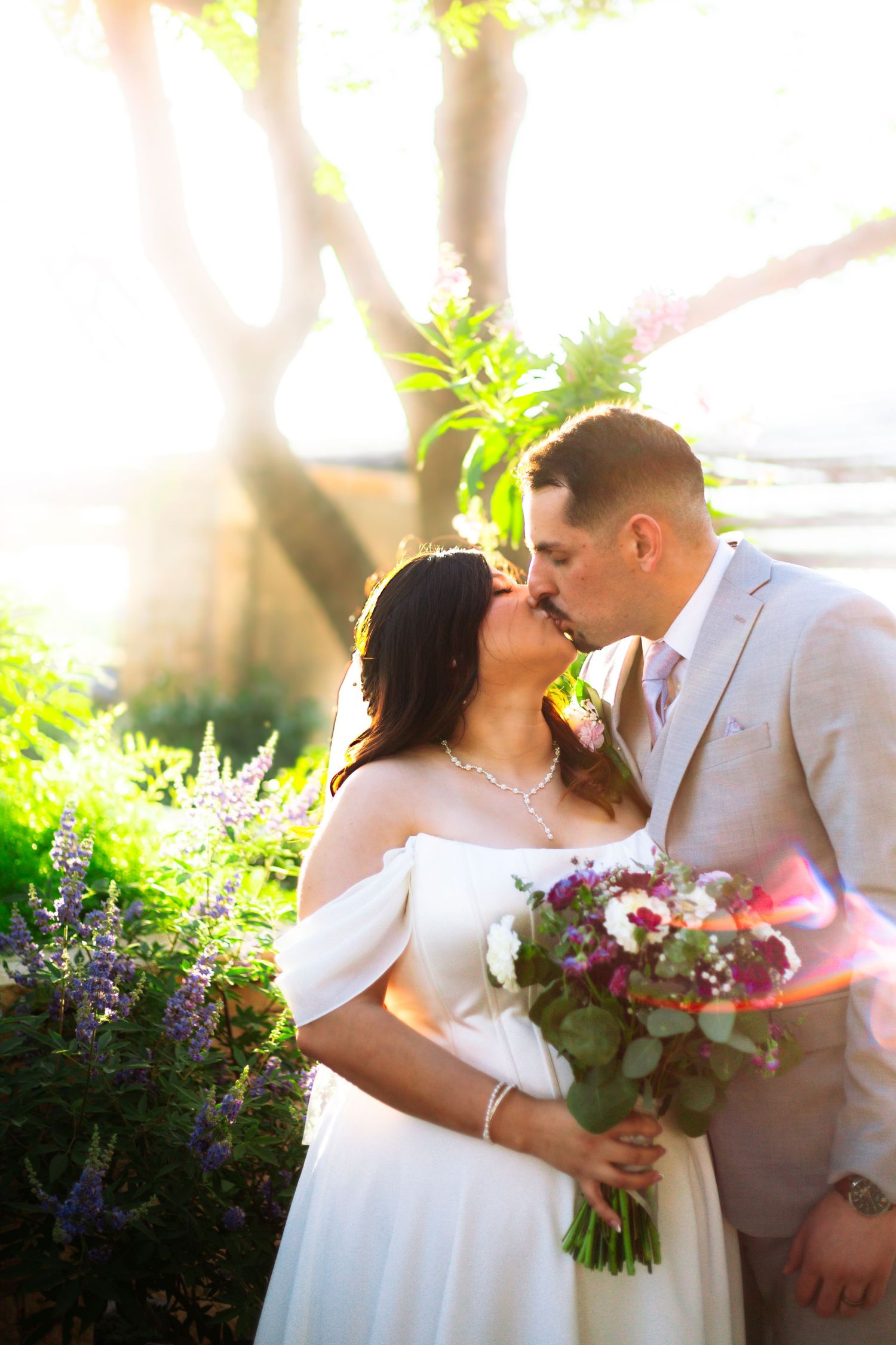 Newlyweds share a romantic kiss outdoors in golden sunlight, bride holding a colorful bridal bouquet.