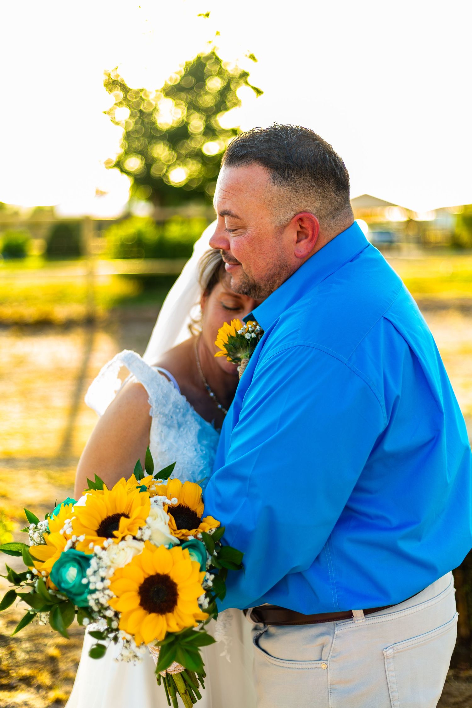 Bride and groom embracing at sunset, bride holding sunflower bouquet, groom in blue shirt, outdoor wedding photo.