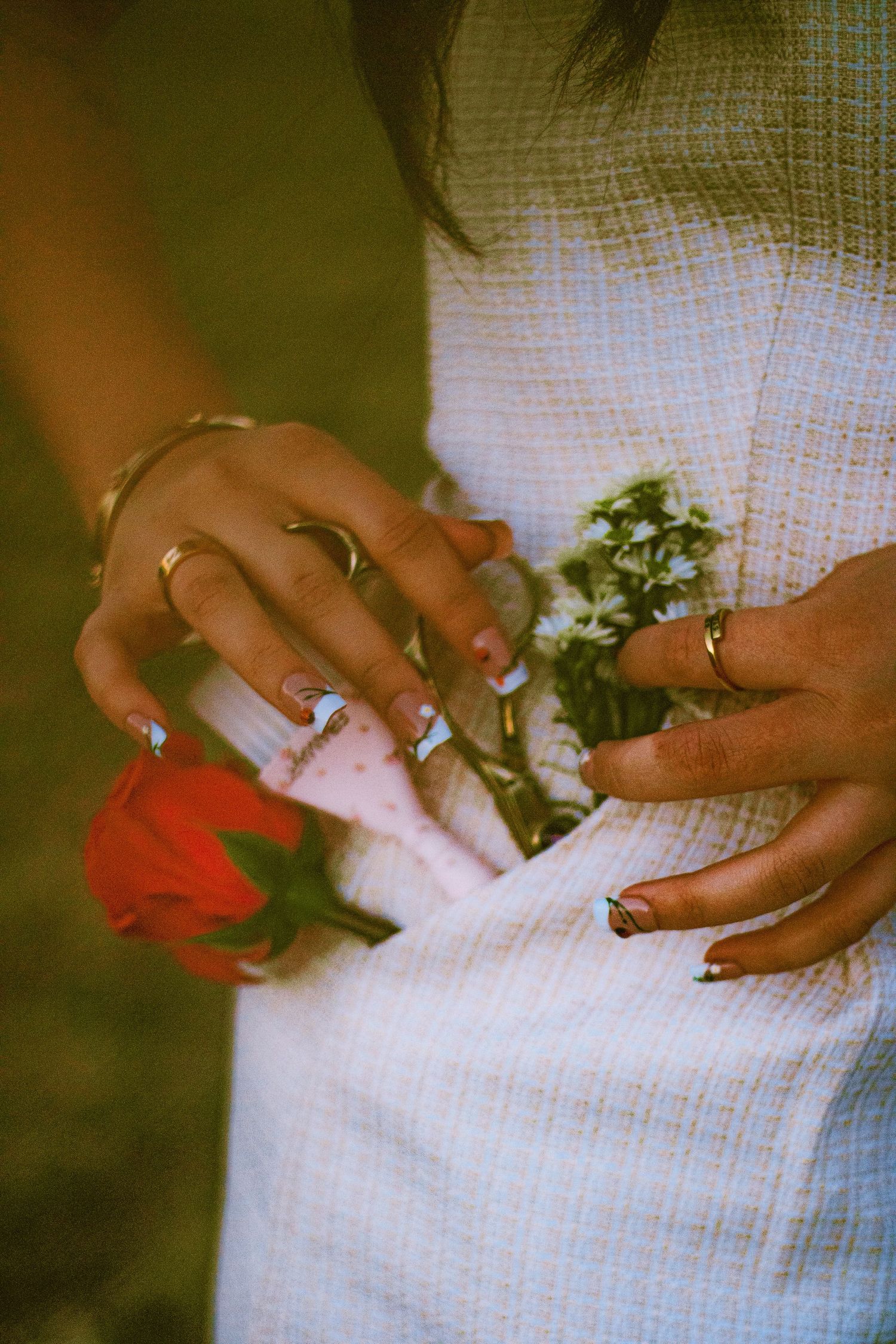 Woman in white dress holding a red rose and white flowers, wearing gold rings and decorated nails.