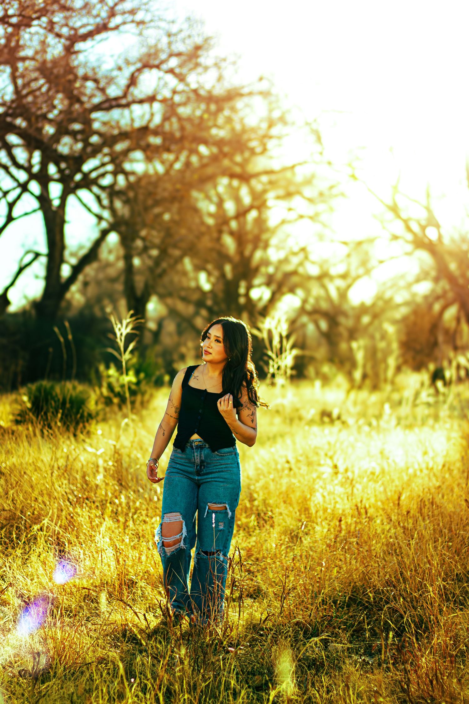 Young woman in black top and ripped jeans standing in golden sunlit field with bare trees in background.