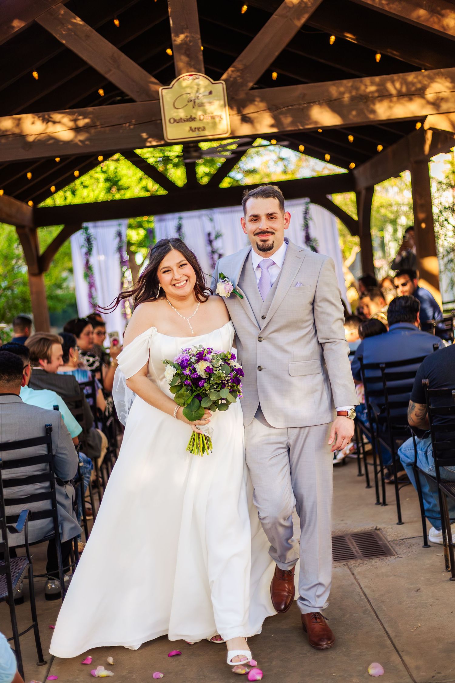 Happy newlyweds walking down the aisle under a wooden gazebo, bride holding purple floral bouquet.