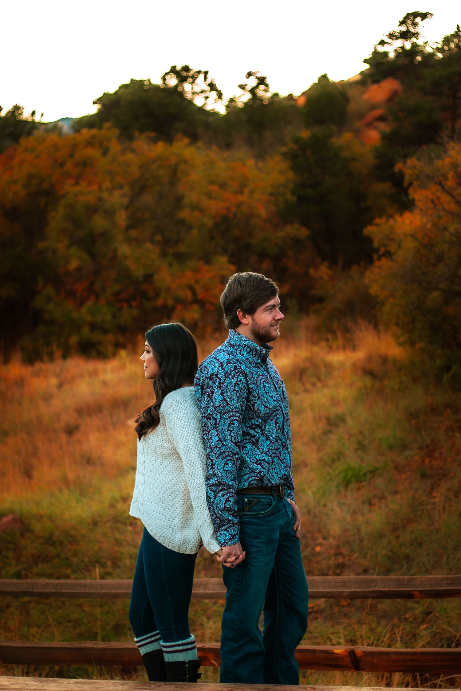 Couple holding hands back-to-back in autumn landscape with colorful fall foliage at sunset.