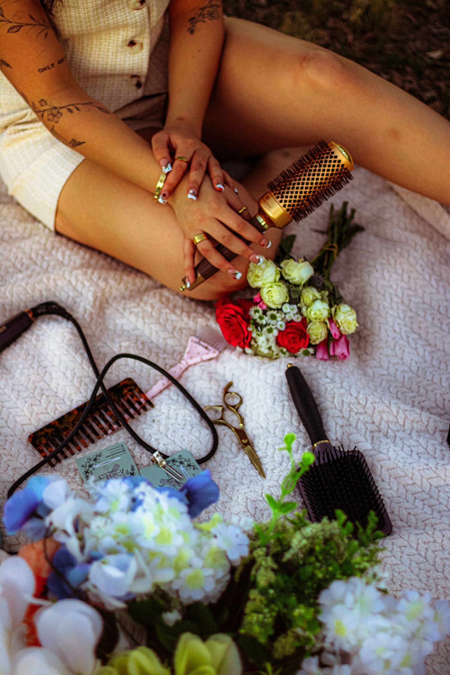 Person with tattooed arms and gold nails holding a round brush, surrounded by hair tools and colorful flowers on a blanket.