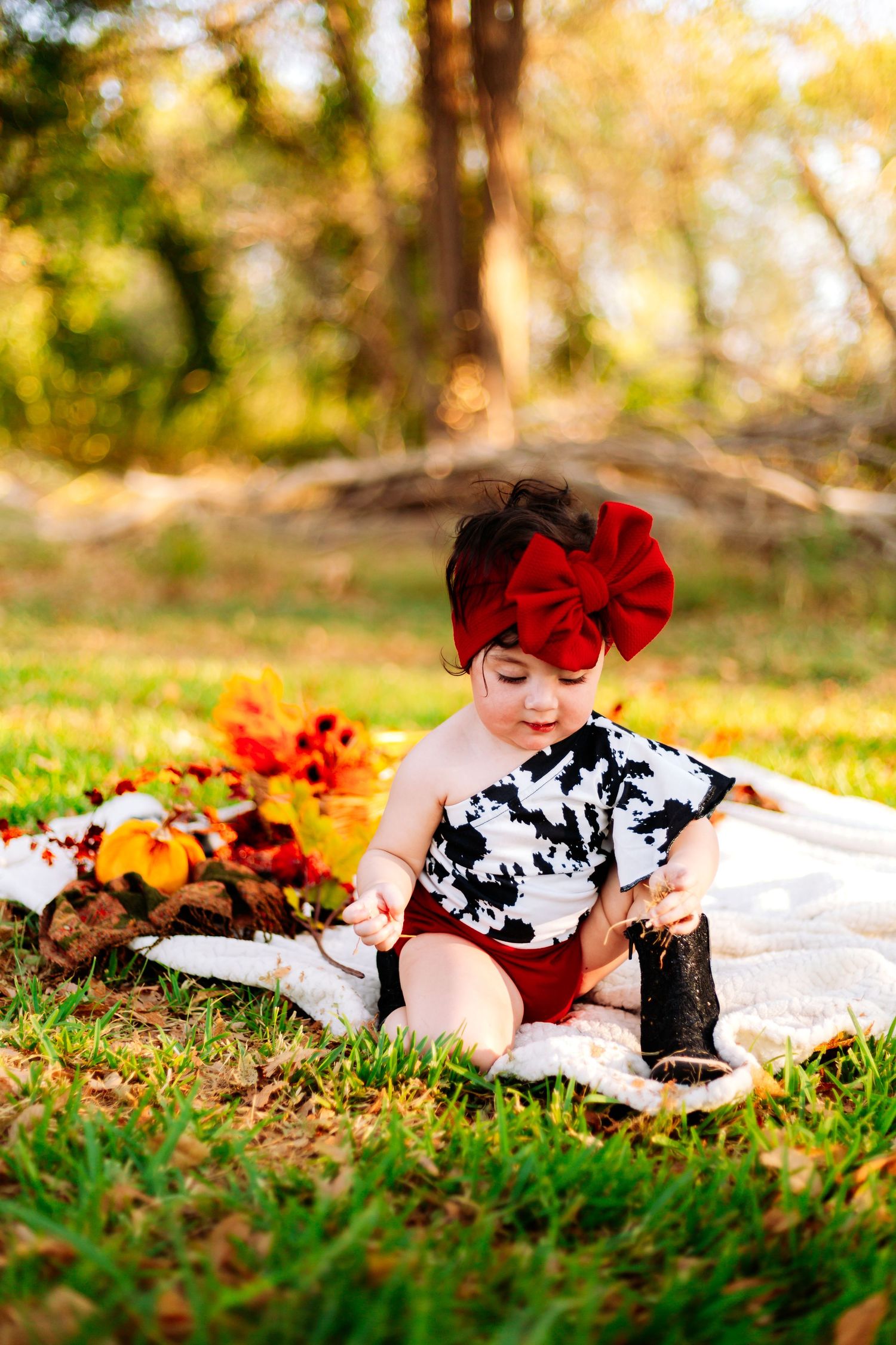 Baby girl in cow print romper and red bow headband sits on blanket with fall leaves outdoors.
