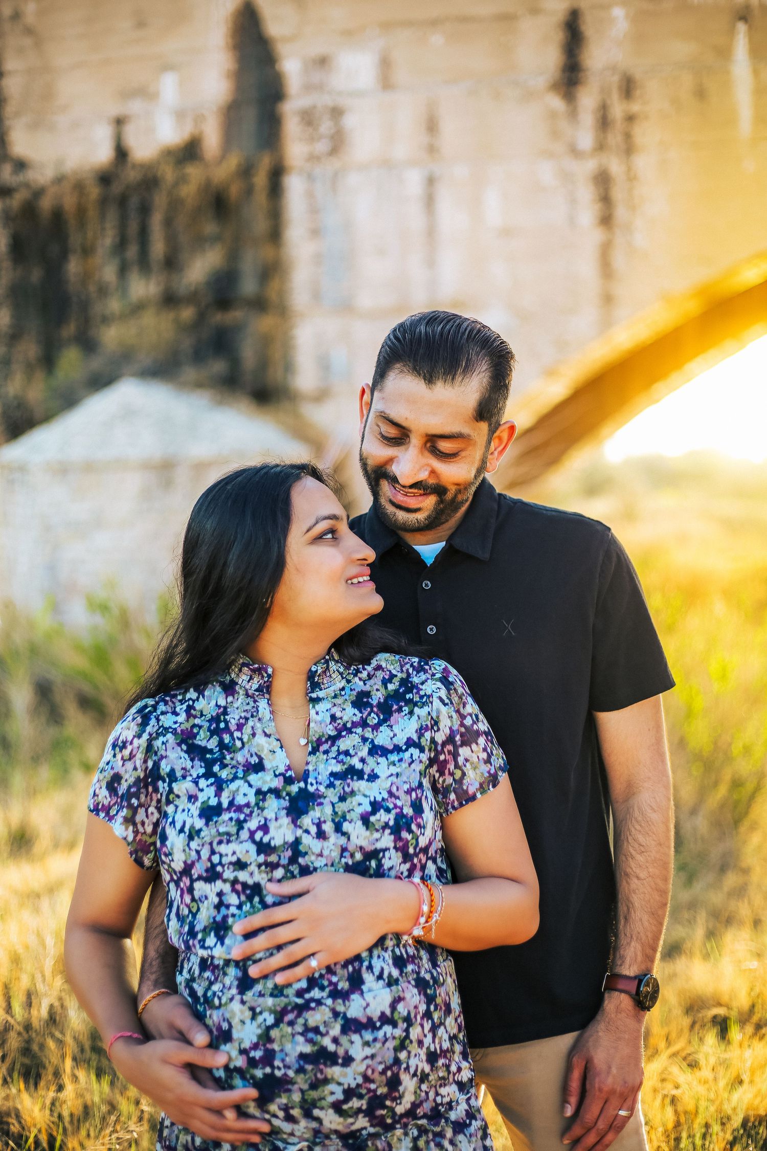 Happy expecting couple embracing at golden hour near rustic concrete wall outdoors.