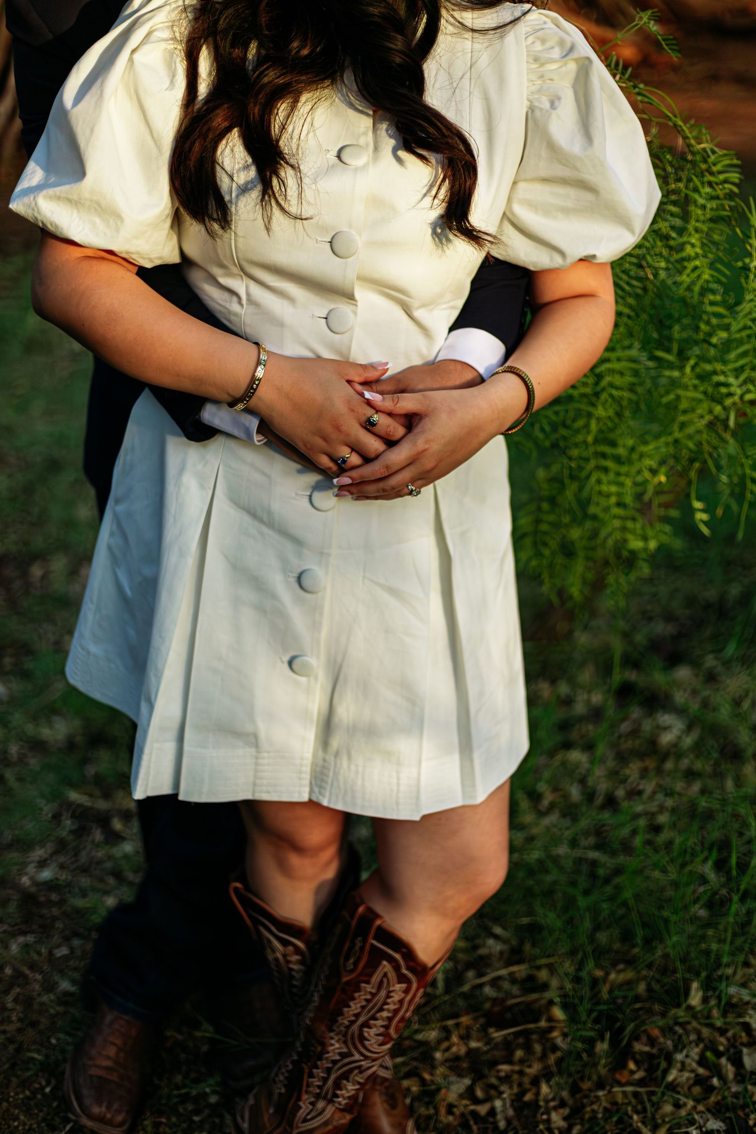Couple embracing outdoors, woman in white button-up dress and brown cowboy boots, hands clasped together.