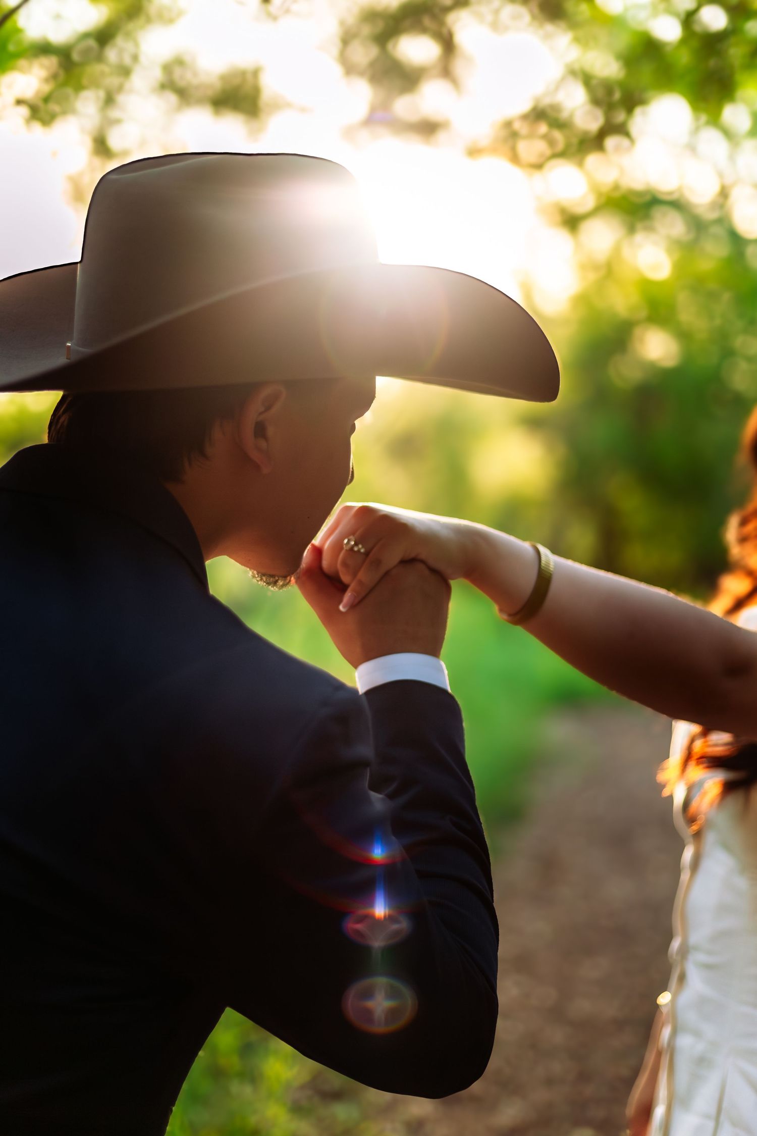 Man in cowboy hat kisses woman's hand at sunset wedding, backlit by golden light in lush green outdoor setting.
